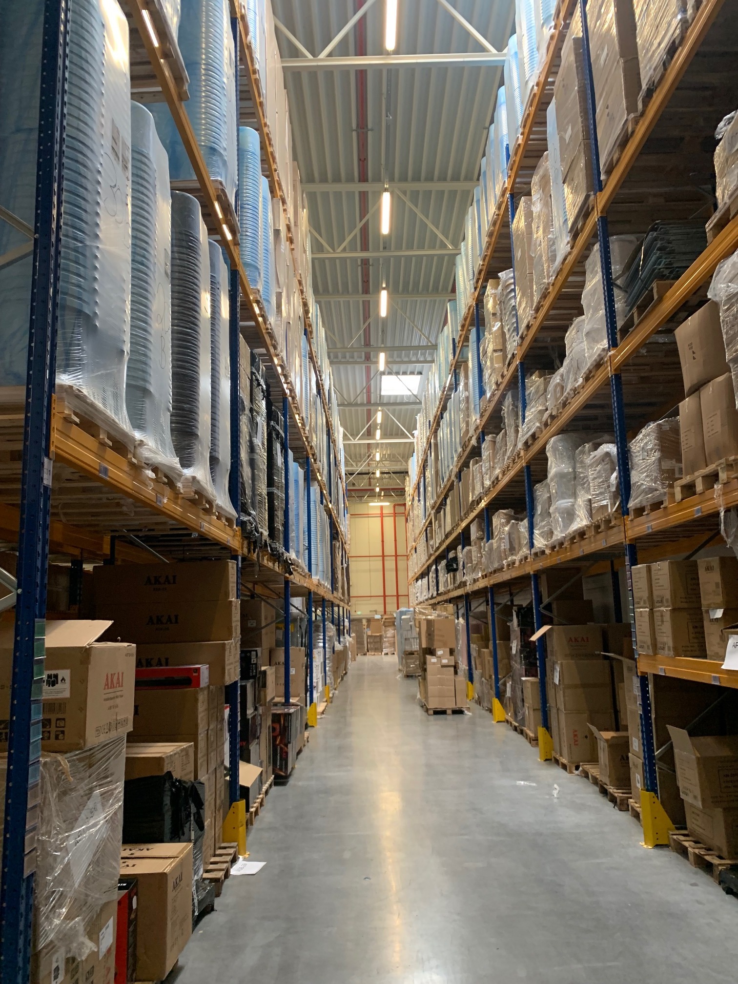 Wnętrze magazynu z wysokimi regałami wypełnionymi kartonami i towarami w folii. Widok w perspektywie wzdłuż korytarza. Metalowa konstrukcja regałów.