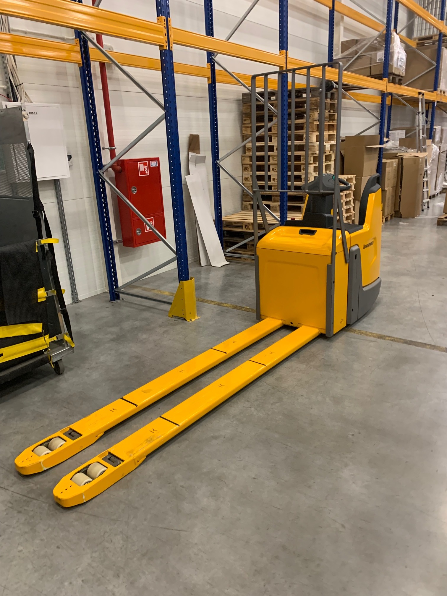 Żółty wózek widłowy Jungheinrich na tle regałów magazynowych z paletami i kartonami, widoczna gaśnica. Magazyn z betonową podłogą.