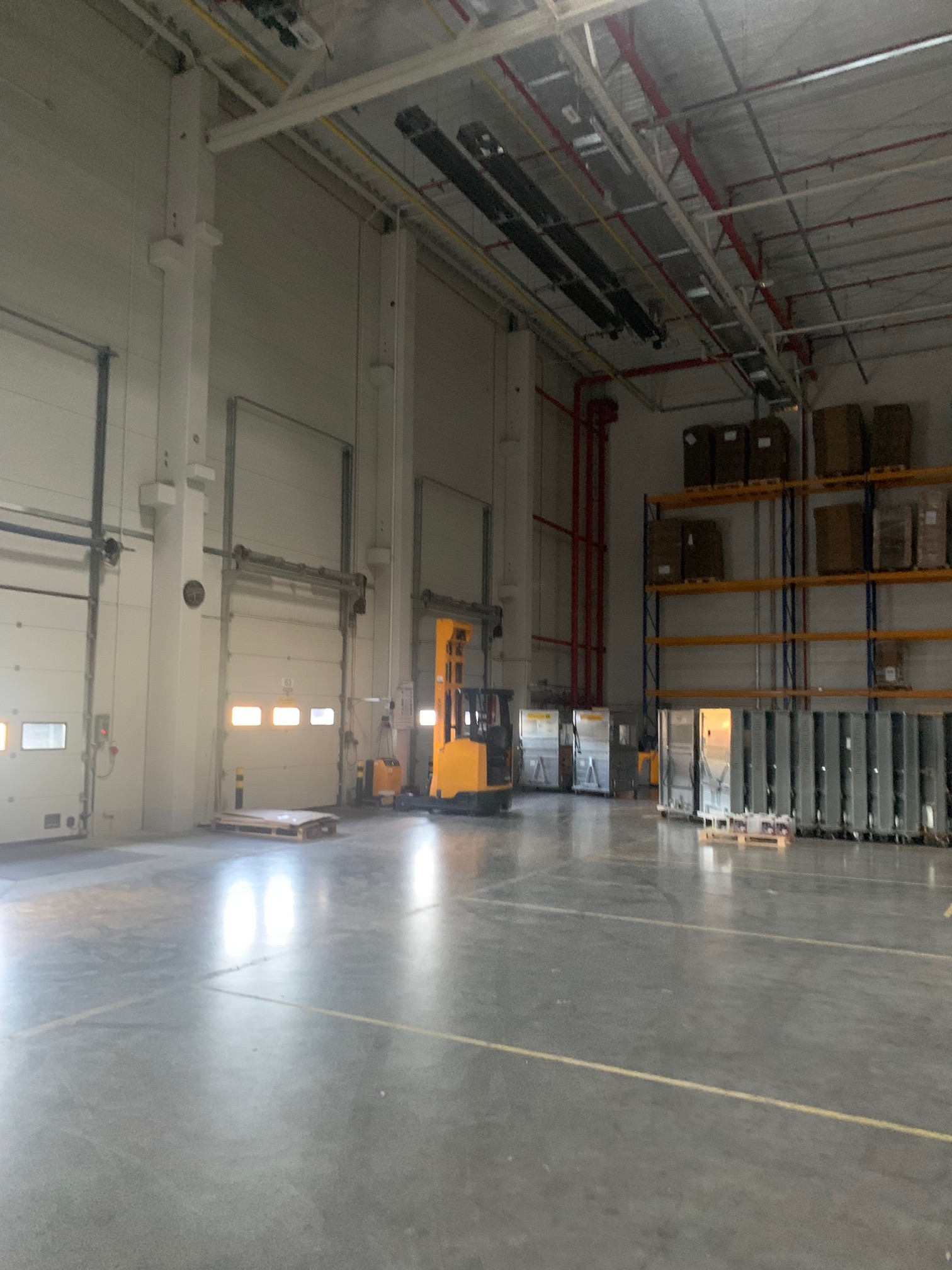 Wnętrze magazynu z regałami, wózkiem widłowym i bramami. Widoczne instalacje pod sufitem i oznaczenia na podłodze. Perspektywa z głębi hali.