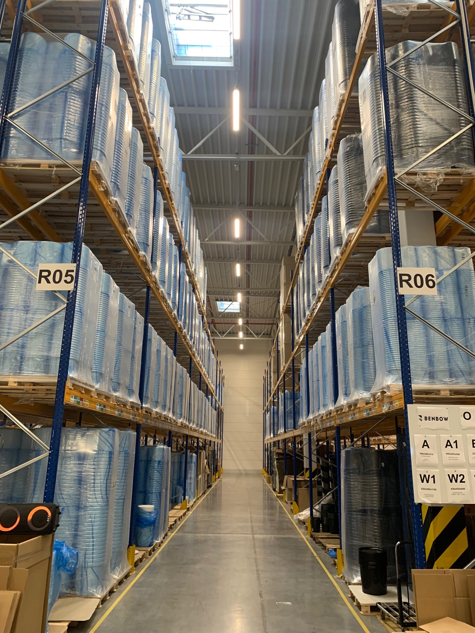 Wysokie regały magazynowe z paletami, na których umieszczono zapakowane pojemniki. Widoczne oznaczenia regałów R05 i R06, oświetlenie sufitowe i betonowa posadzka.