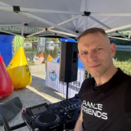 DJ na zewnątrz, obok bojek Aqua Speed i nagłośnienia, na tle jeziora podczas imprezy sportowej. Widoczny sprzęt Pioneer DJ i baner 24h Water Open Swimming.