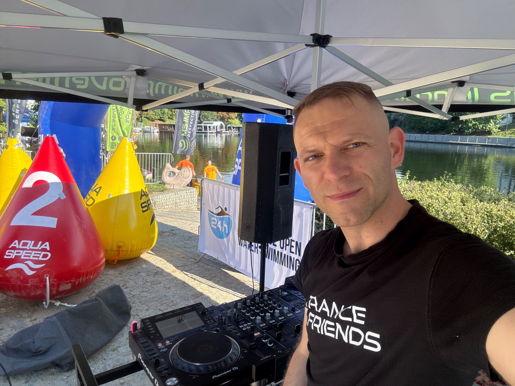 DJ na zewnątrz, obok bojek Aqua Speed i nagłośnienia, na tle jeziora podczas imprezy sportowej. Widoczny sprzęt Pioneer DJ i baner 24h Water Open Swimming.