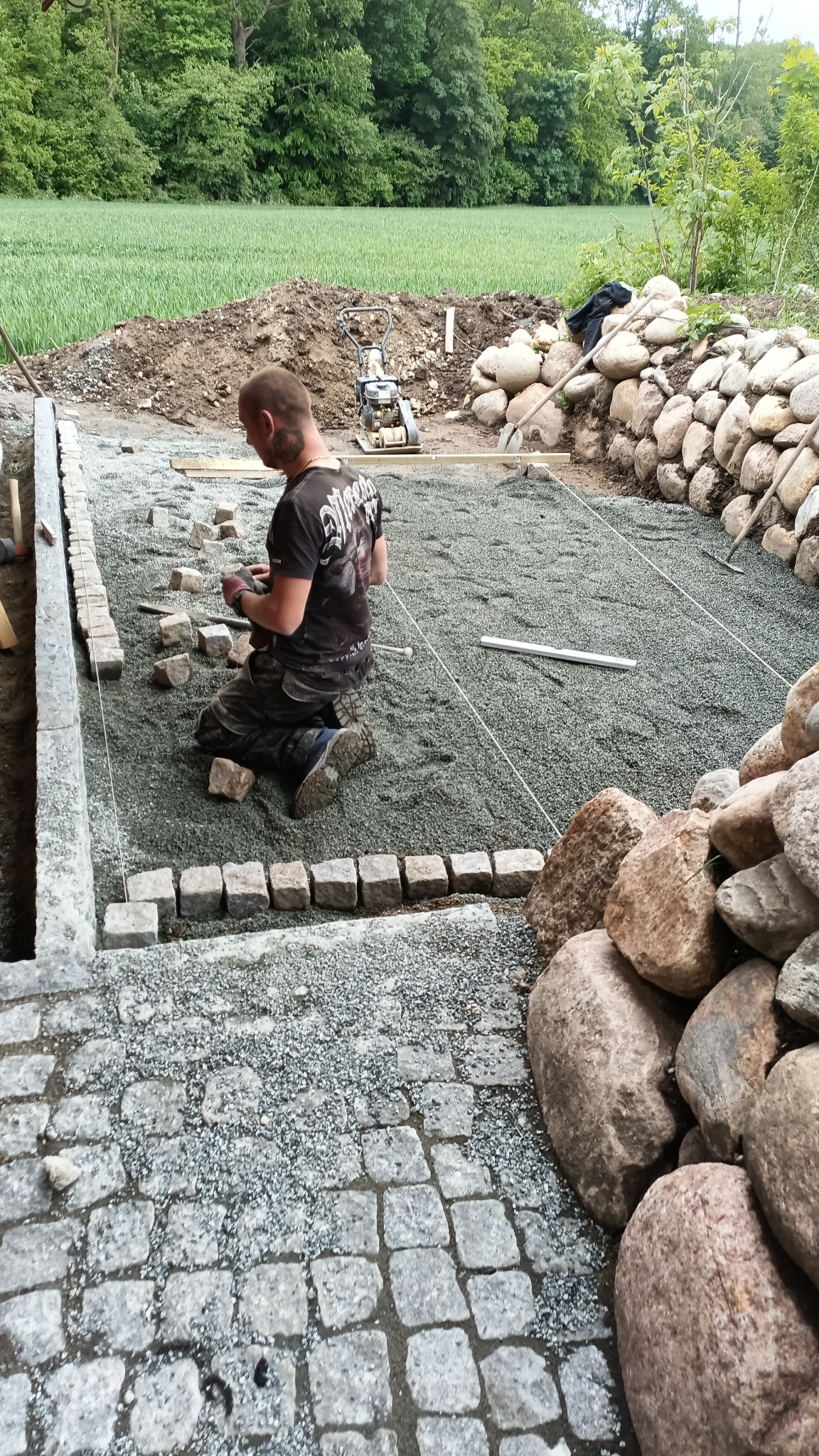 Układanie kostki granitowej: brukarz klęczy, układając obrzeża z kostki. W tle ubijarka, przygotowany teren i kamienny murek. Widoczny postęp prac.