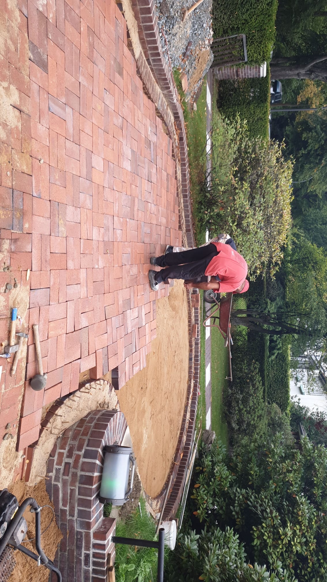 Układanie czerwonej kostki brukowej w jodełkę, z obrzeżem z cegły. Widoczny etap prac z piaskiem, narzędzia brukarskie i pracownik w czerwonej koszulce.