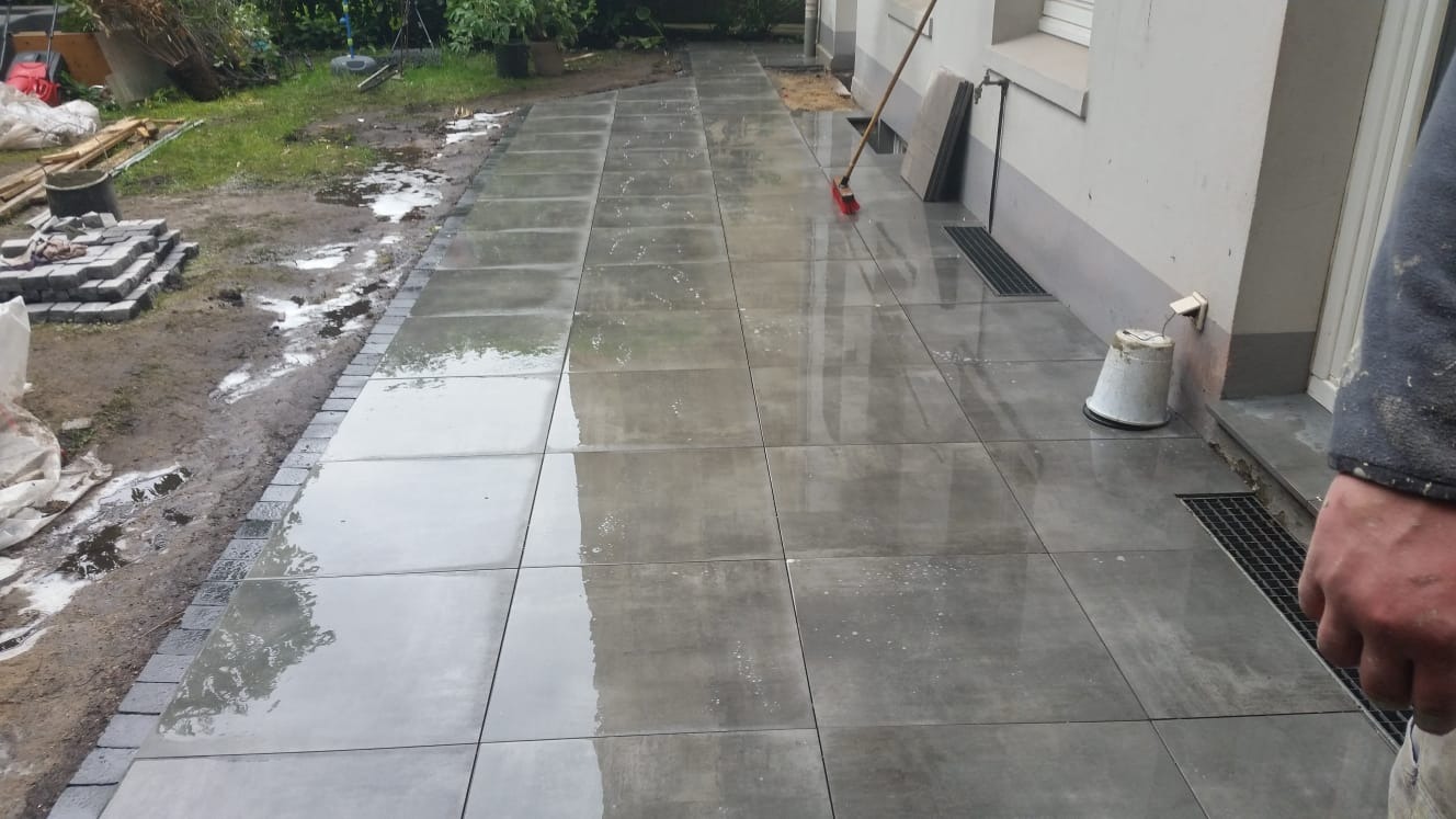 Świeżo ułożona, mokra, szara kostka brukowa na posesji, z widocznym obrzeżem i stertą kostki w tle. Odbicia światła na powierzchni płytek.