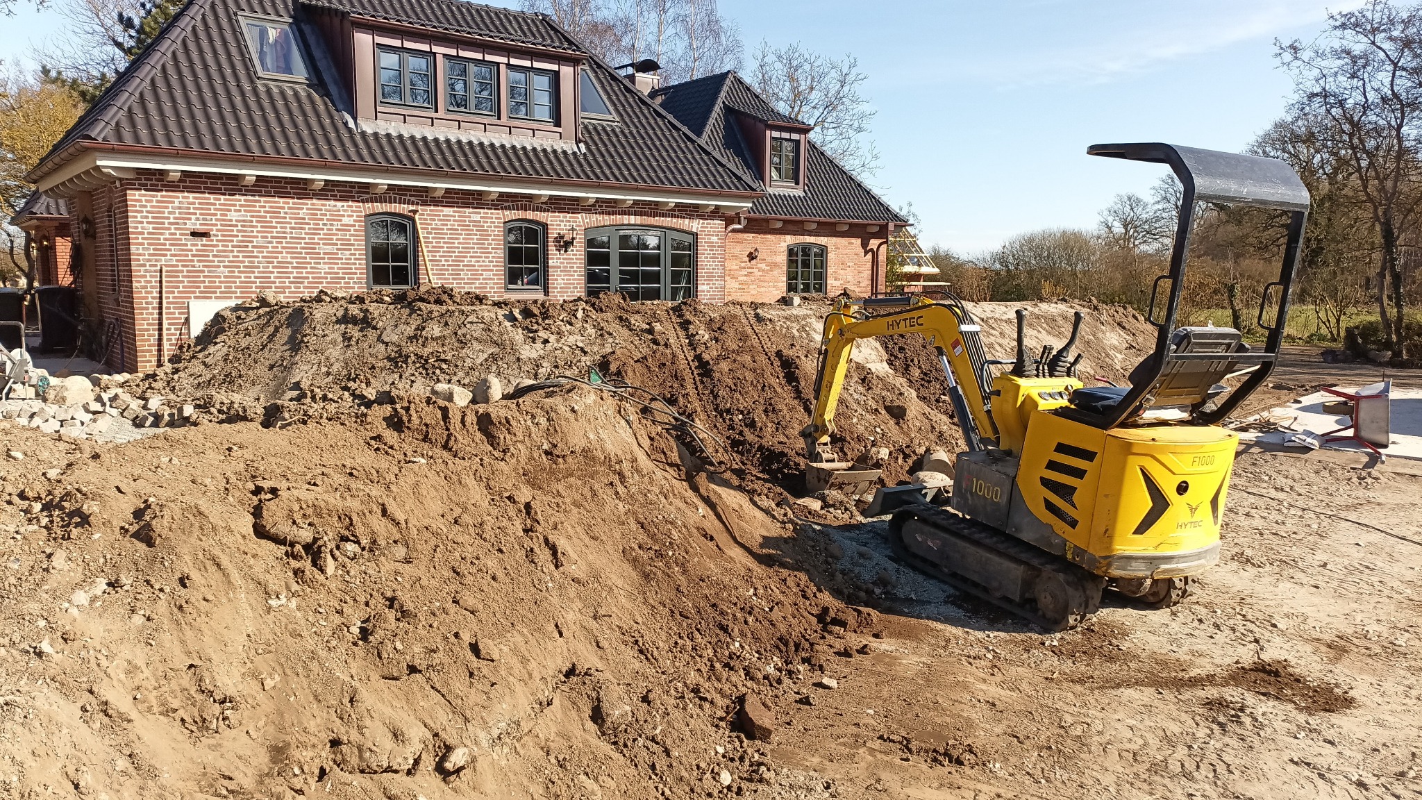 Żółta minikoparka Hytec 1000 na tle wykopu ziemnego przed ceglanym domem z ciemną dachówką. Prace ziemne w toku, słoneczny dzień.