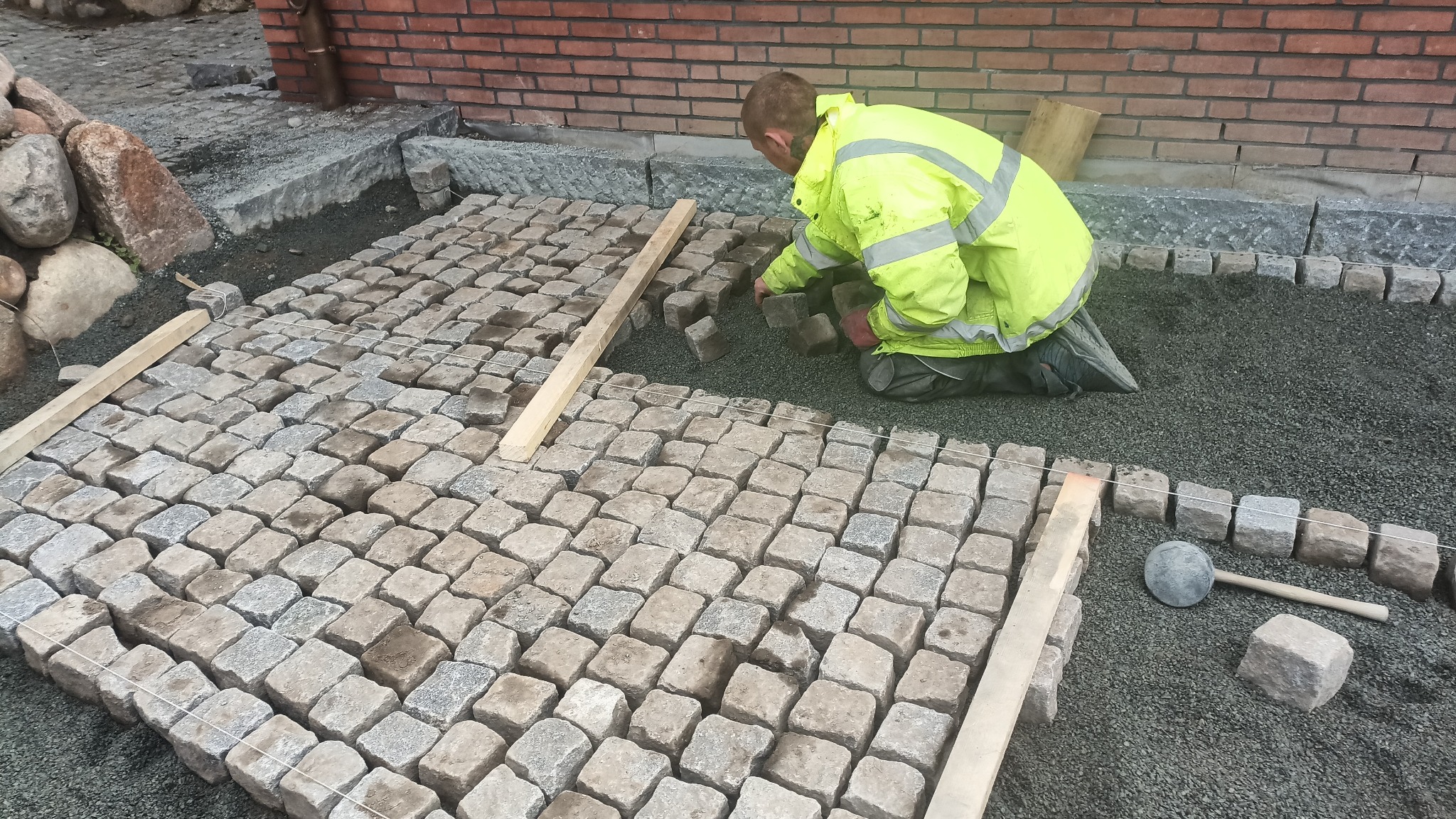 Układanie kostki granitowej: brukarz w żółtej kurtce układa kostkę na podsypce, widoczne drewniane listwy i gumowy młotek. Fragment muru z cegły w tle.
