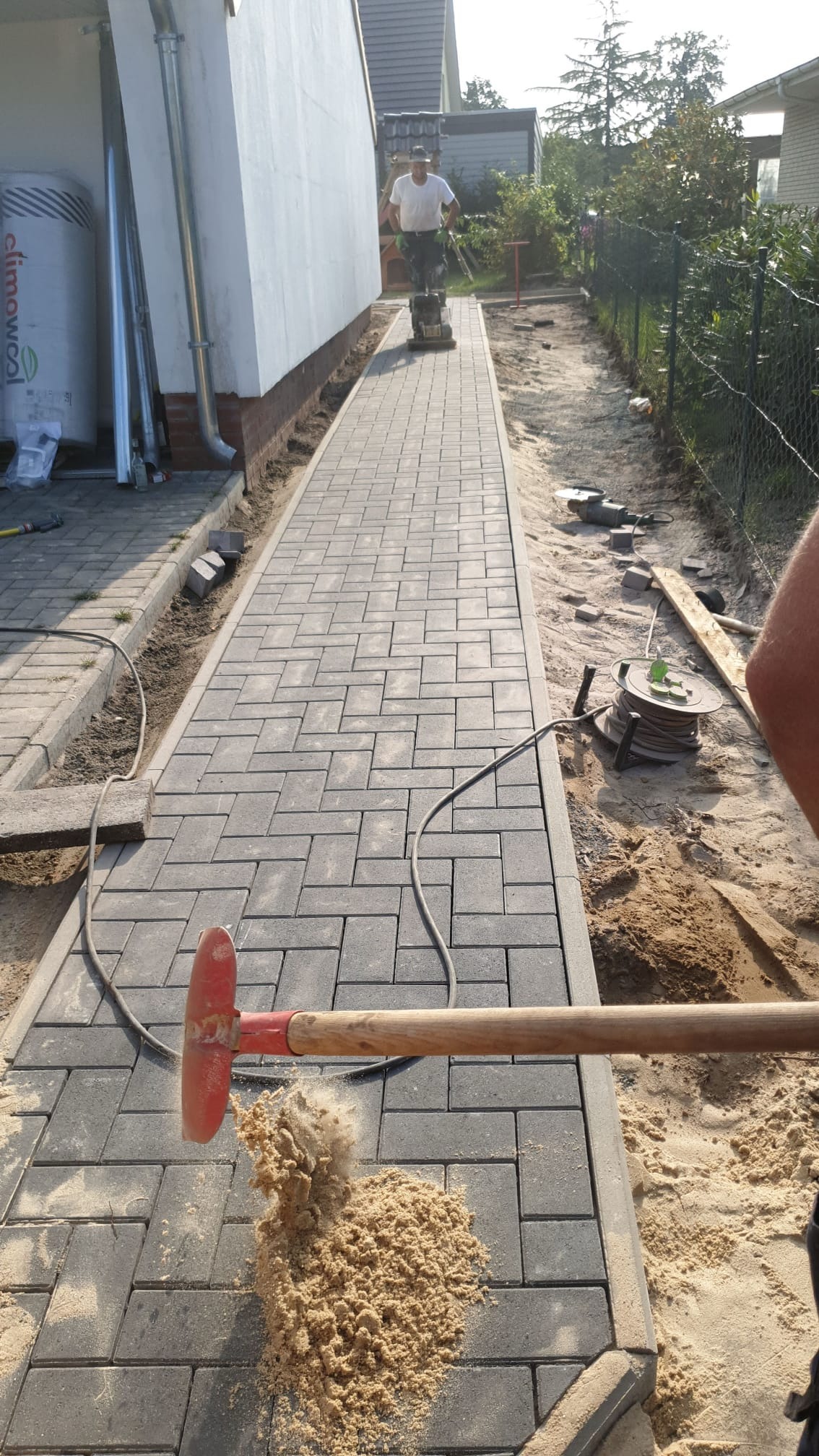 Układanie szarej kostki brukowej na posesji w Hamburgu. Widoczny pracownik z ubijarką i narzędzia brukarskie. Kostka ułożona w zygzak, piasek do wypełnienia szczelin.