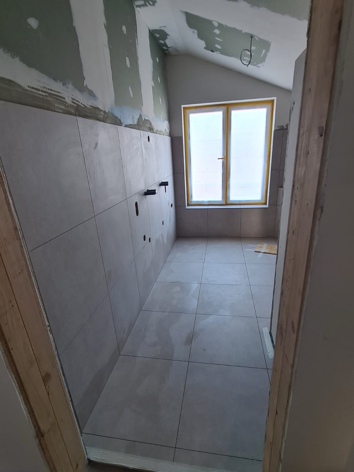 Remont łazienki na poddaszu: ułożone szare płytki na ścianach i podłodze, przygotowane instalacje. Okno z żółtą ramą. Ściany częściowo w gładzi.