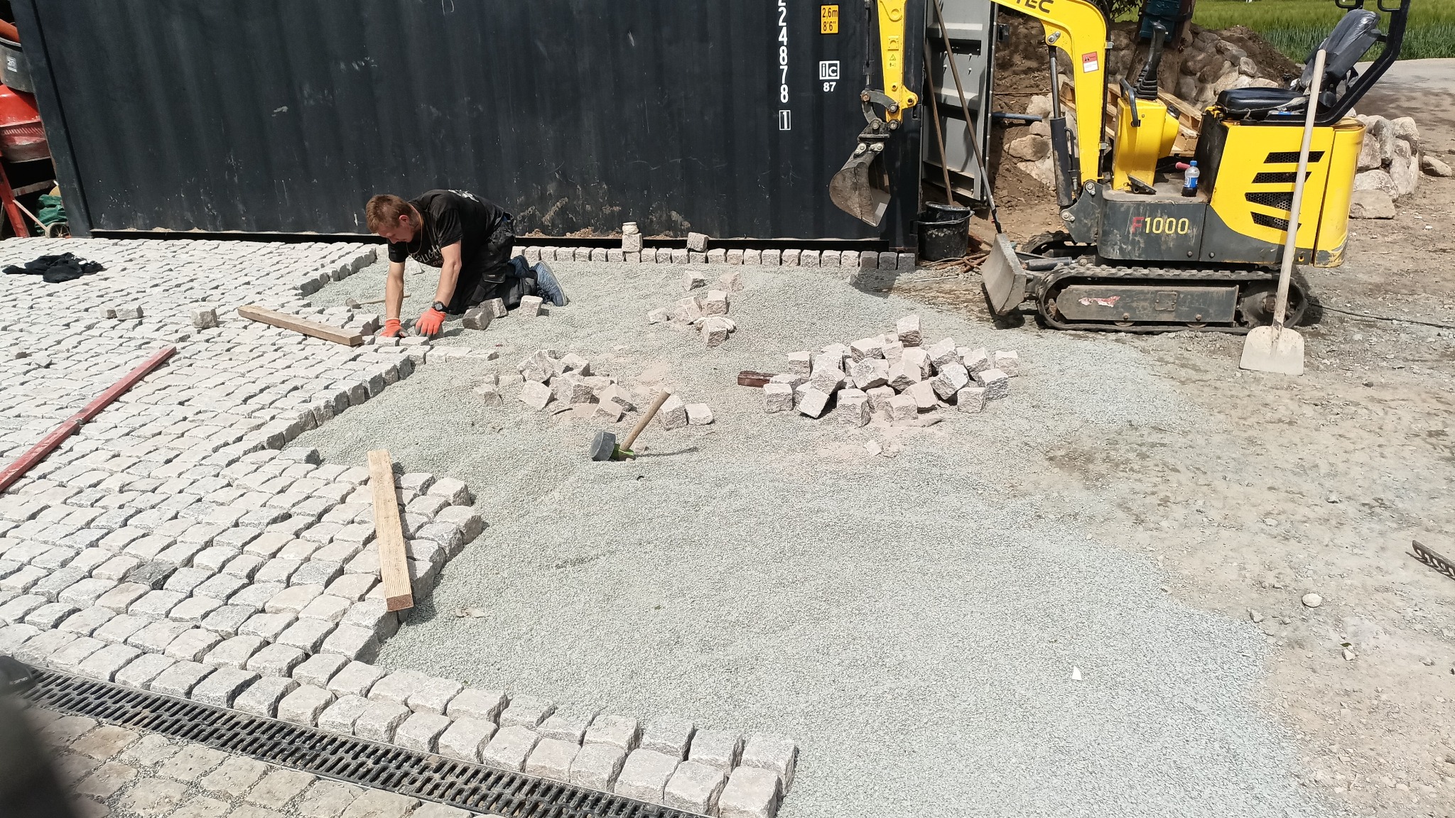 Układanie kostki granitowej: mężczyzna w pomarańczowych rękawicach, kostka, podsypka, minikoparka TEC F1000. Widok z góry na plac budowy.