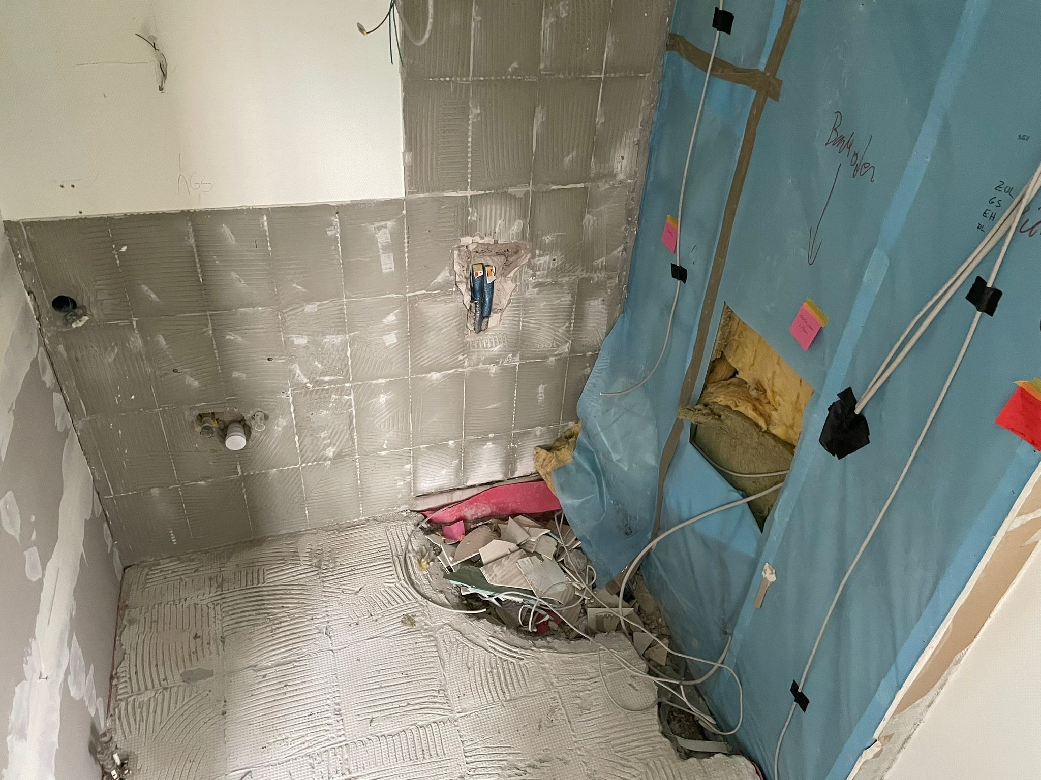 Wnętrze łazienki w trakcie remontu: położone płytki, instalacje, zabezpieczone ściany, widoczne przewody i gruz. Etap prac wykończeniowych.