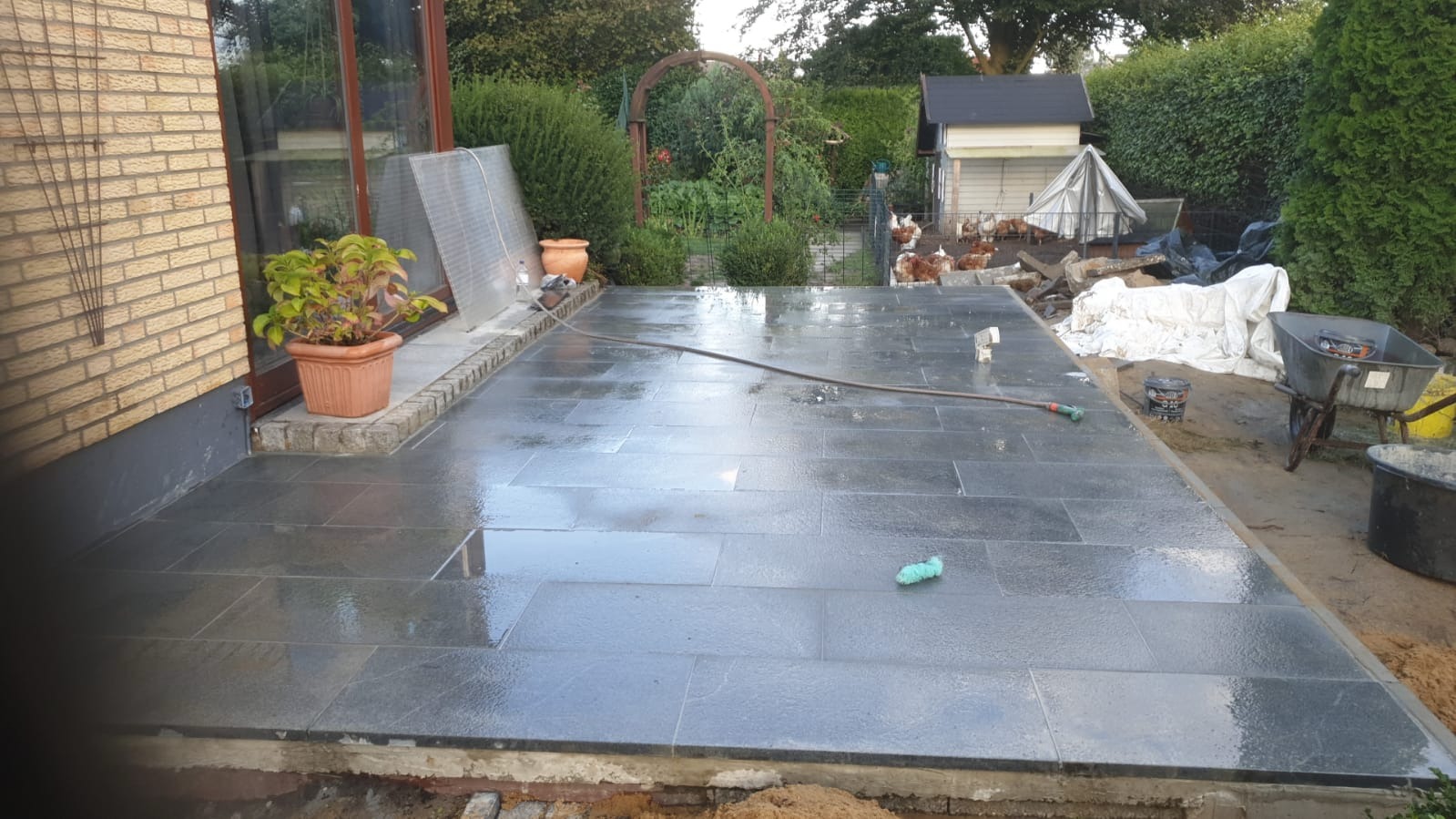 Świeżo ułożona, mokra, ciemnoszara kostka brukowa tworzy taras. W tle ogród z pergolą, budynkiem gospodarczym i kurami. Część kostki jest ucięta w kadrze.