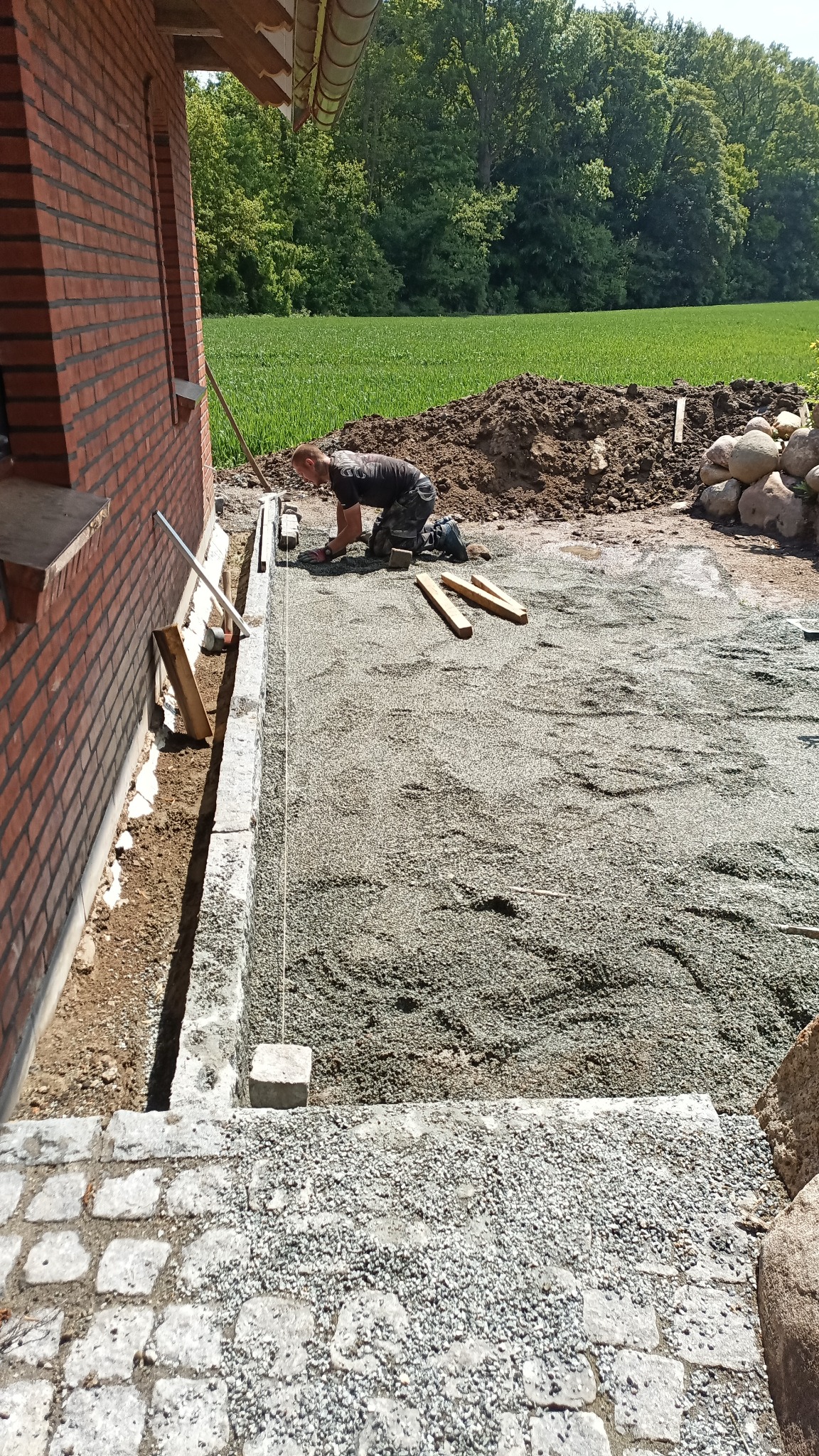 Mężczyzna na kolanach układa kostkę brukową na podsypce przy budynku z cegły, widoczne narzędzia i materiały budowlane, w tle zielone pole i drzewa.