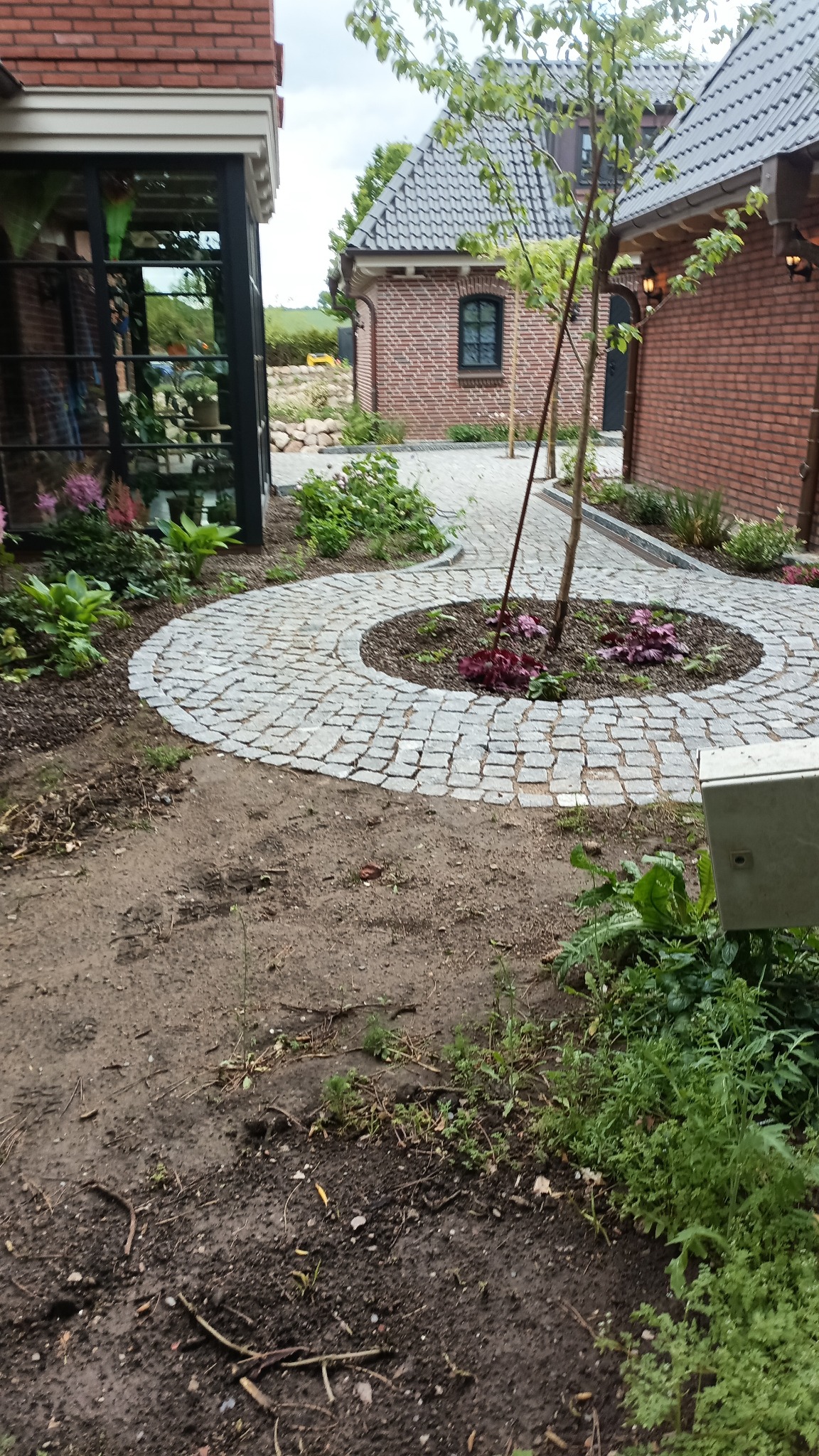 Okrągły chodnik z kostki granitowej otaczający rabatę z drzewem i roślinami, prowadzący do budynku z cegły. Widoczne elementy architektury krajobrazu.