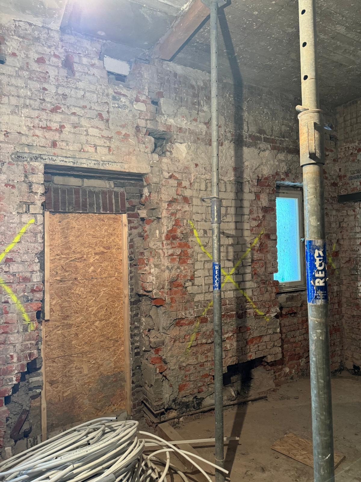 Ceglana ściana w trakcie rozbiórki, z widocznymi śladami po usuniętych fragmentach, zabezpieczone otwory, metalowe podpory i zwój kabli na podłodze.