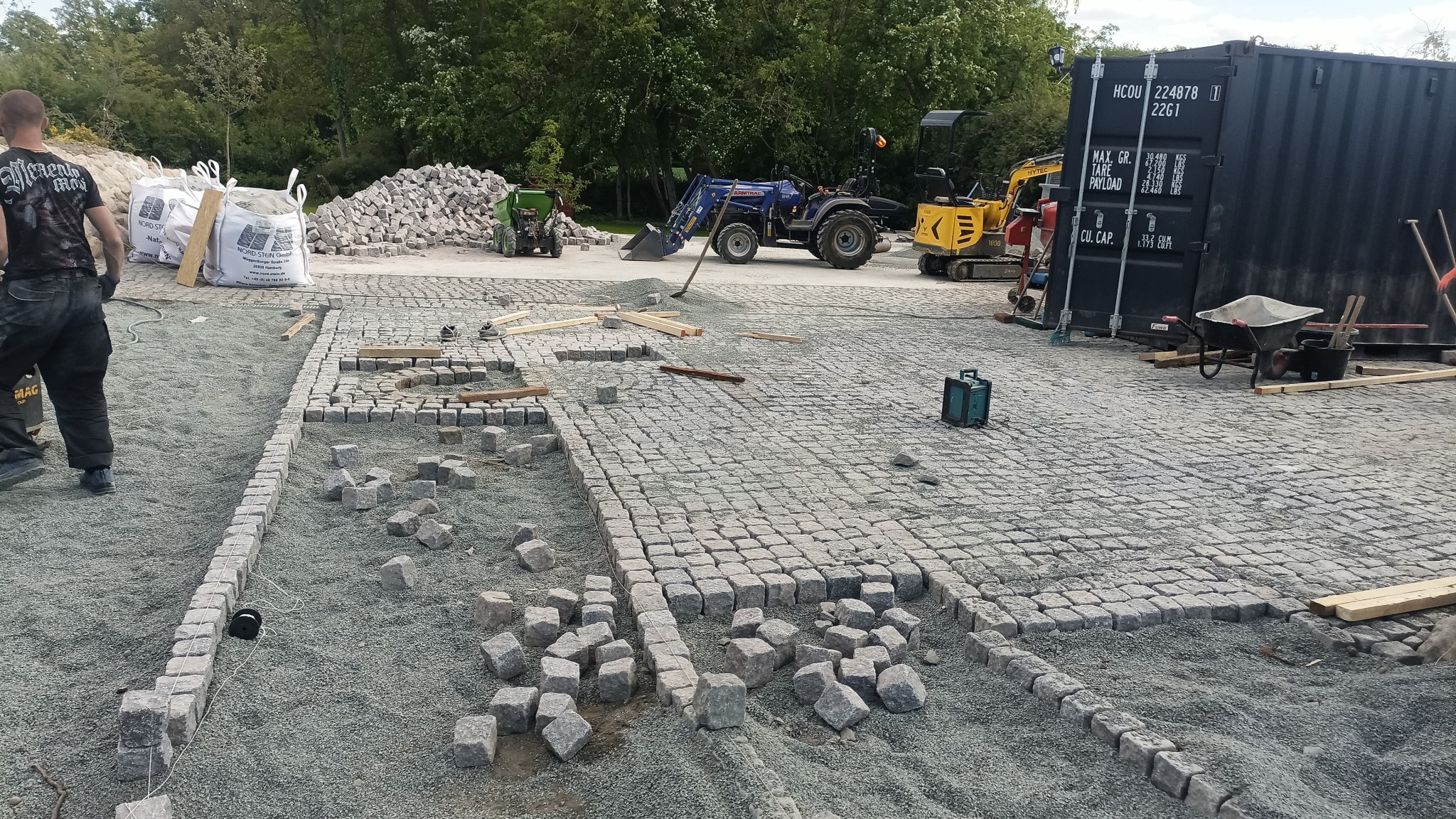 Układanie kostki granitowej na podsypce, widoczne maszyny budowlane i pracownik. Częściowo ułożona nawierzchnia z kostki, worki z materiałem budowlanym w tle.