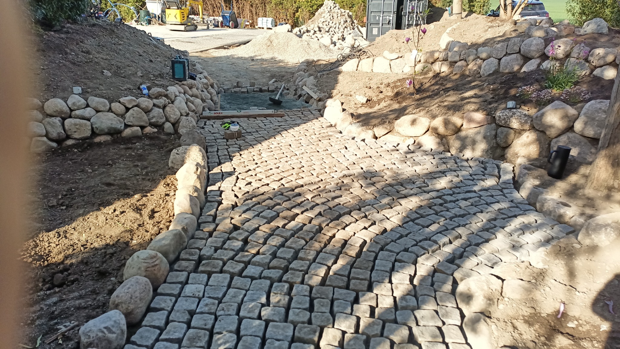 Układanie kostki granitowej w ogrodzie: ścieżka z kostki, obrzeża z kamieni, wykopana ziemia, narzędzia pracy, koparka w tle. Etap prac w słoneczny dzień.