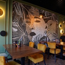 BUDART EXPERT ARTEM SKLIAR - Mozaika z motywem twarzy i roślin na ścianie w lokalu. Oświetlenie punktowe, drewniane stoły i żółte krzesła tworzą przytulną atmosferę.