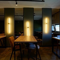 BUDART EXPERT ARTEM SKLIAR - Nowoczesne oświetlenie ścienne w lokalu usługowym. Pionowe lampy z okrągłym akcentem na tle paneli i luster. Widoczne stoły i krzesła w tle.