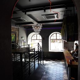 BUDART EXPERT ARTEM SKLIAR - Stylowe wnętrze lokalu z elementami cegły, nowoczesnym oświetleniem i heksagonalną posadzką. Widoczne schody, bar i osoba za nim. Całość utrzymana w ciemnej tonacji.