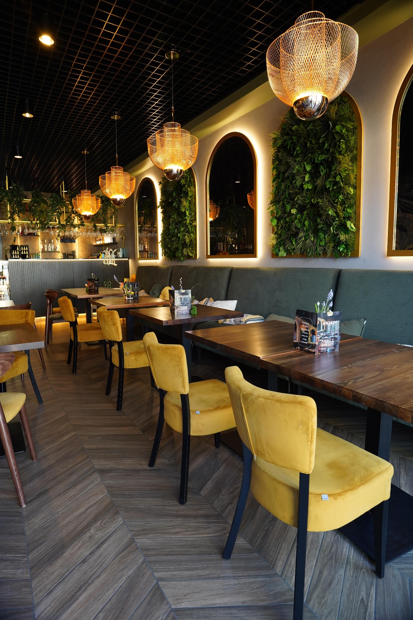 Stylowe wnętrze restauracji z drewnianymi stołami, żółtymi krzesłami i dekoracjami roślinnymi na ścianach. Oświetlenie stanowią ażurowe lampy wiszące.