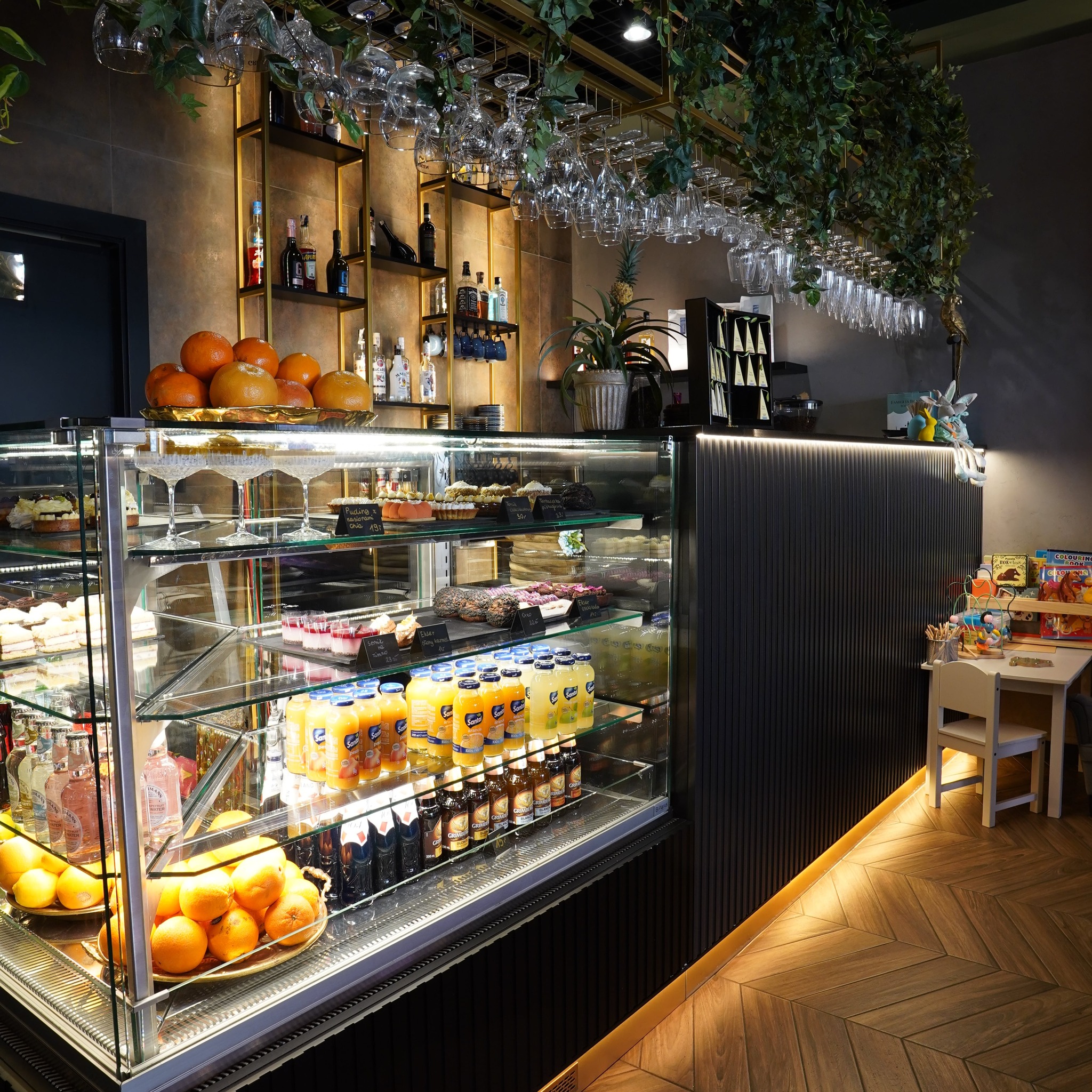 Eleganckie wnętrze kawiarni z przeszkloną ladą pełną deserów, soków i napojów. Na półkach butelki alkoholi, dekoracje roślinne i oświetlenie punktowe.