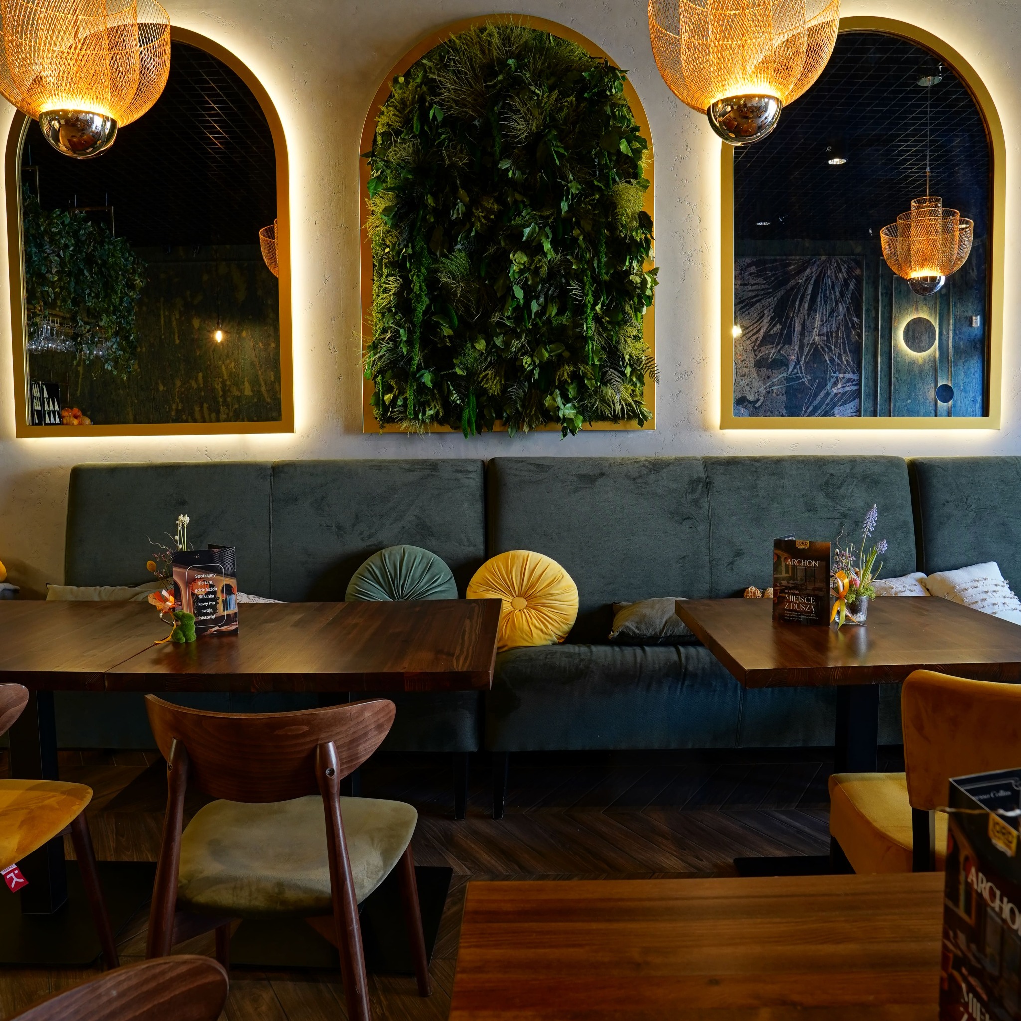 Stylowe wnętrze kawiarni z zieloną ścianą, lustrami w złotych ramach i drewnianymi stolikami. Ciepłe oświetlenie i ciemnozielona sofa tworzą przytulną atmosferę.
