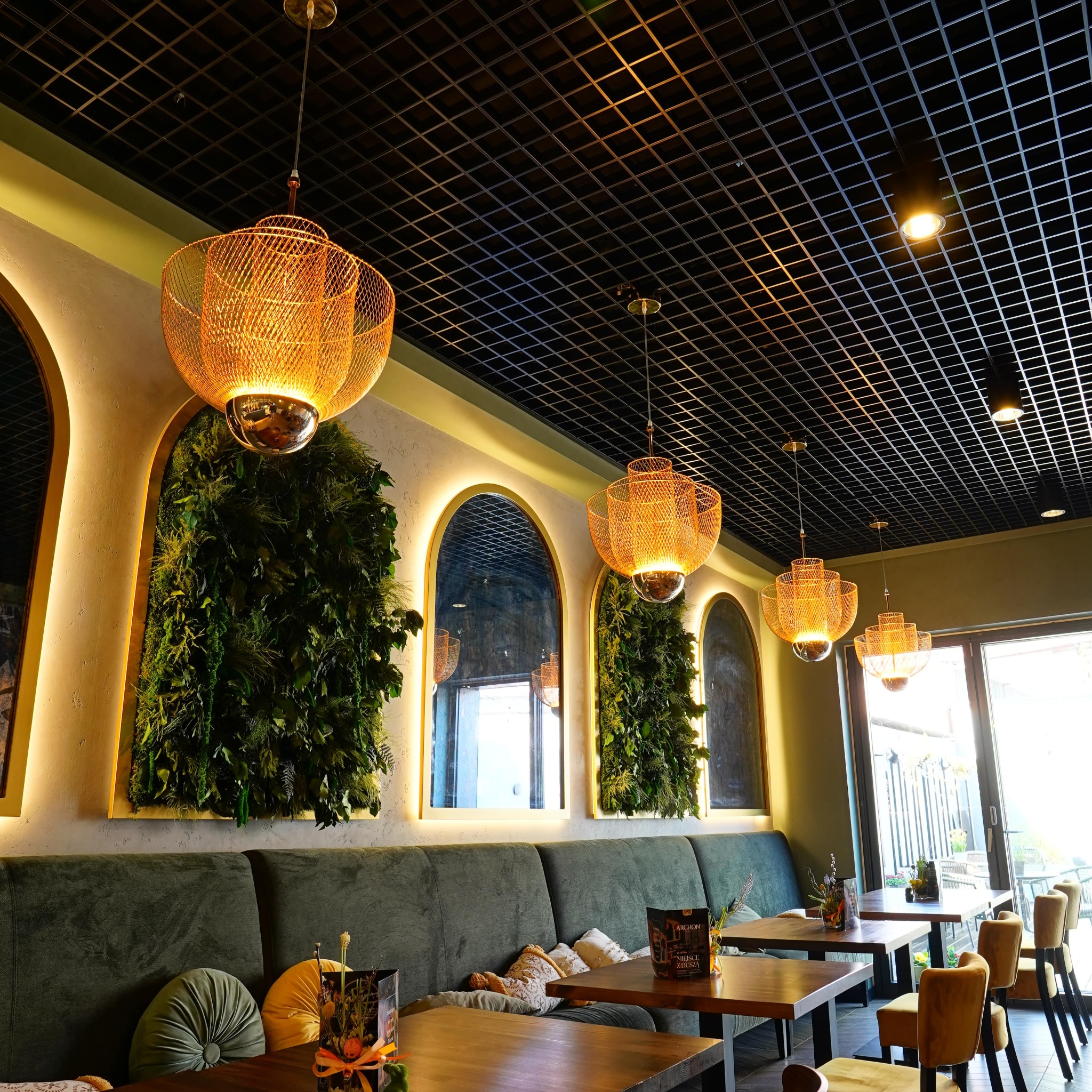 Stylowe wnętrze restauracji z zieloną ścianą, lustrami w złotych ramach i dekoracyjnymi lampami. Widoczny ciemny sufit z kratką i drewniane stoły.