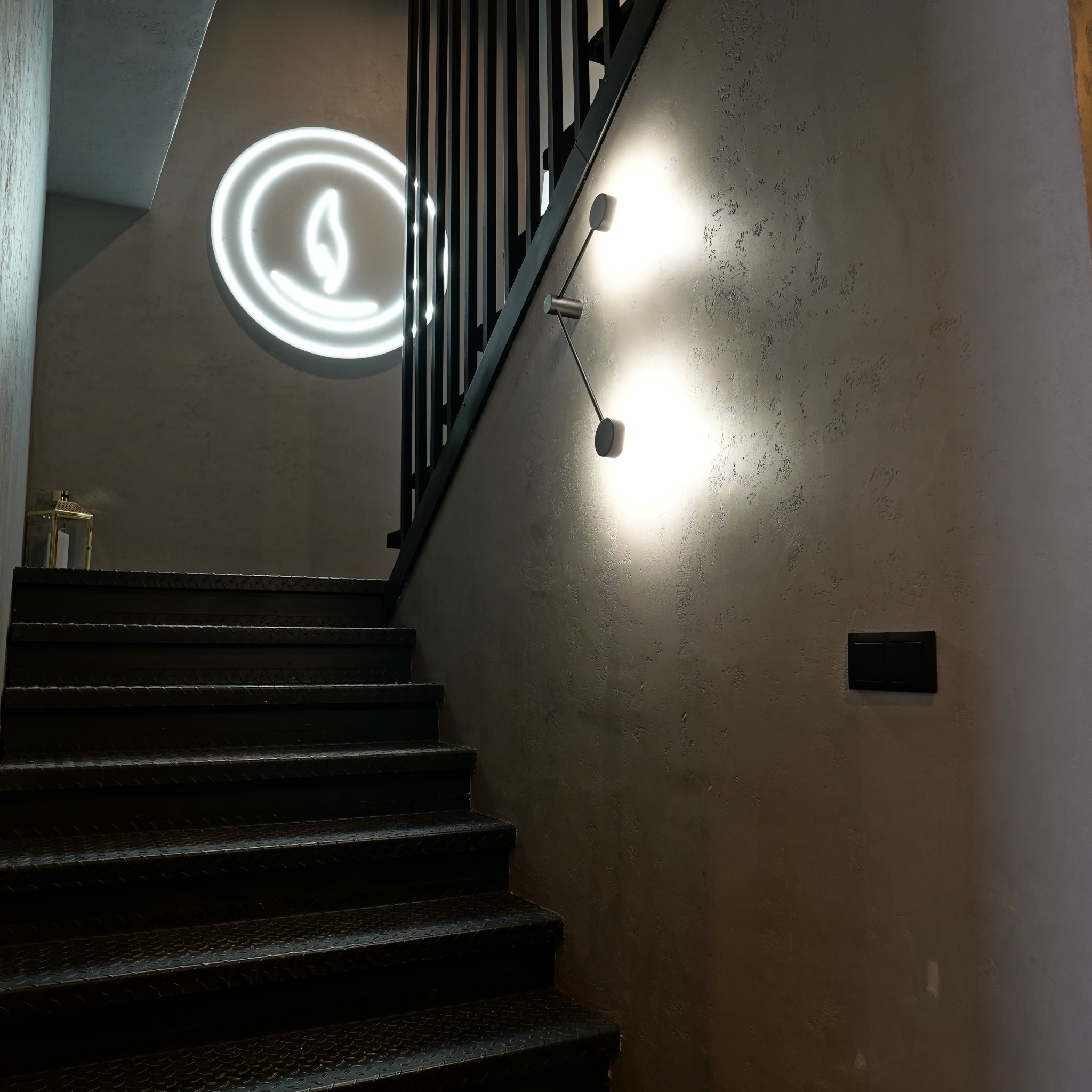 Stylowe schody z metalowymi stopniami i czarną balustradą. Na ścianie lampa i neon w kształcie płomienia. Dekoracyjna faktura ściany.