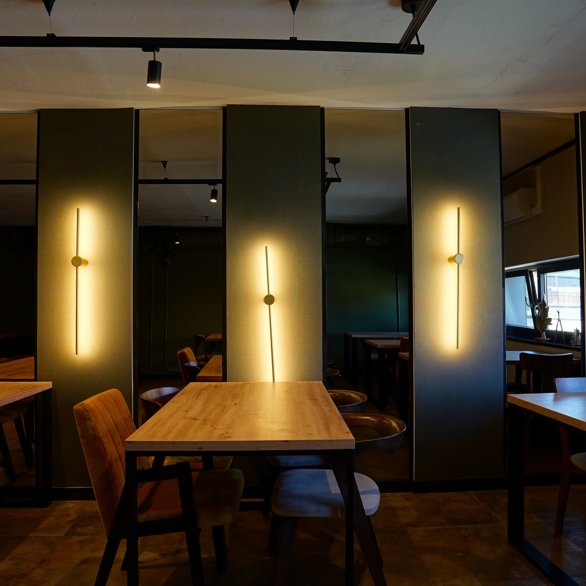 Nowoczesne oświetlenie ścienne w lokalu gastronomicznym. Pionowe lampy LED na tle paneli z lustrami, odbijające wnętrze restauracji. Drewniane stoły i krzesła w ciepłej tonacji.