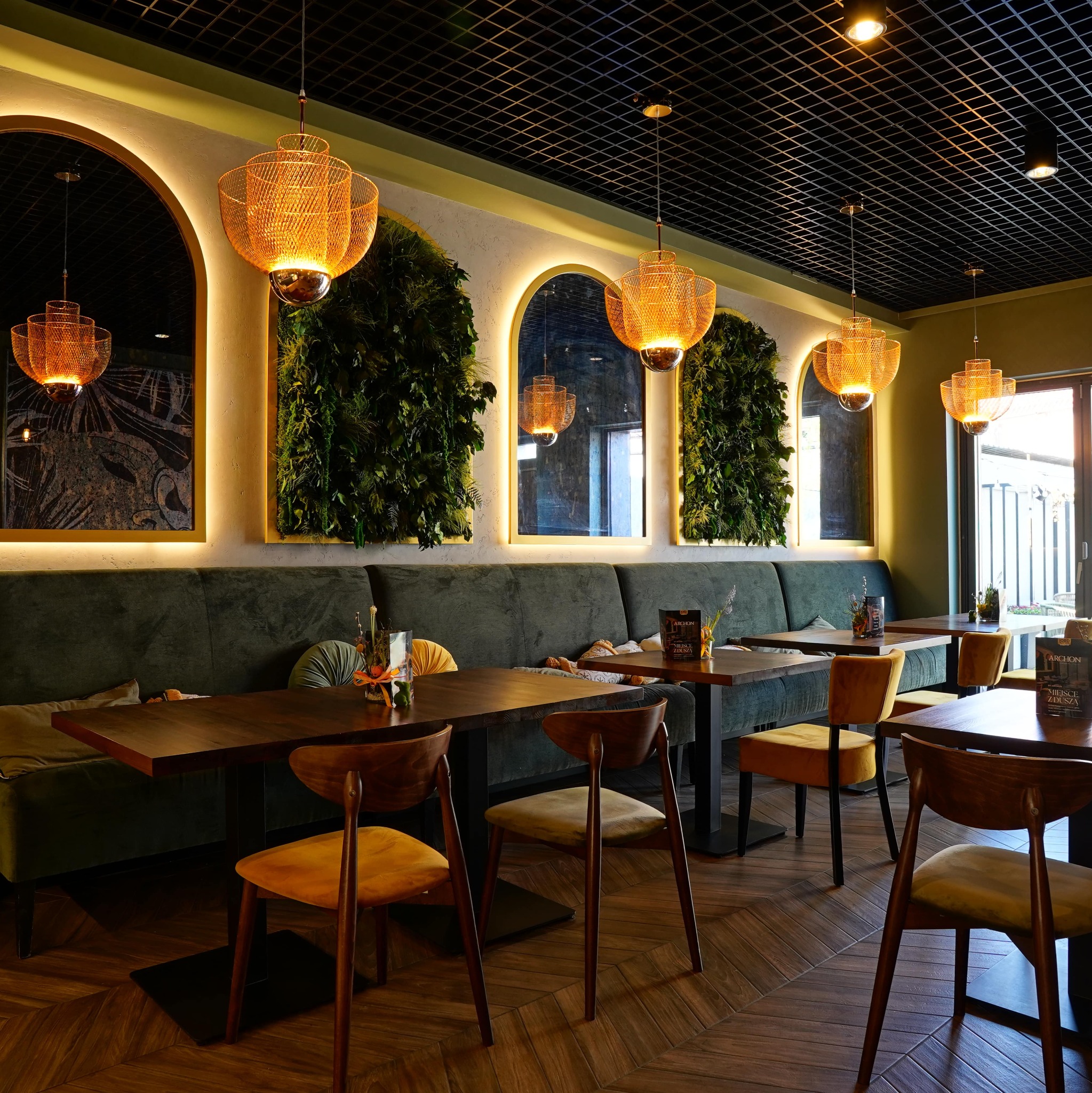 Stylowe wnętrze restauracji z zieloną ścianą, drewnianymi stołami i krzesłami oraz dekoracyjnymi lampami. Widoczny parkiet w jodełkę i podświetlane lustra.
