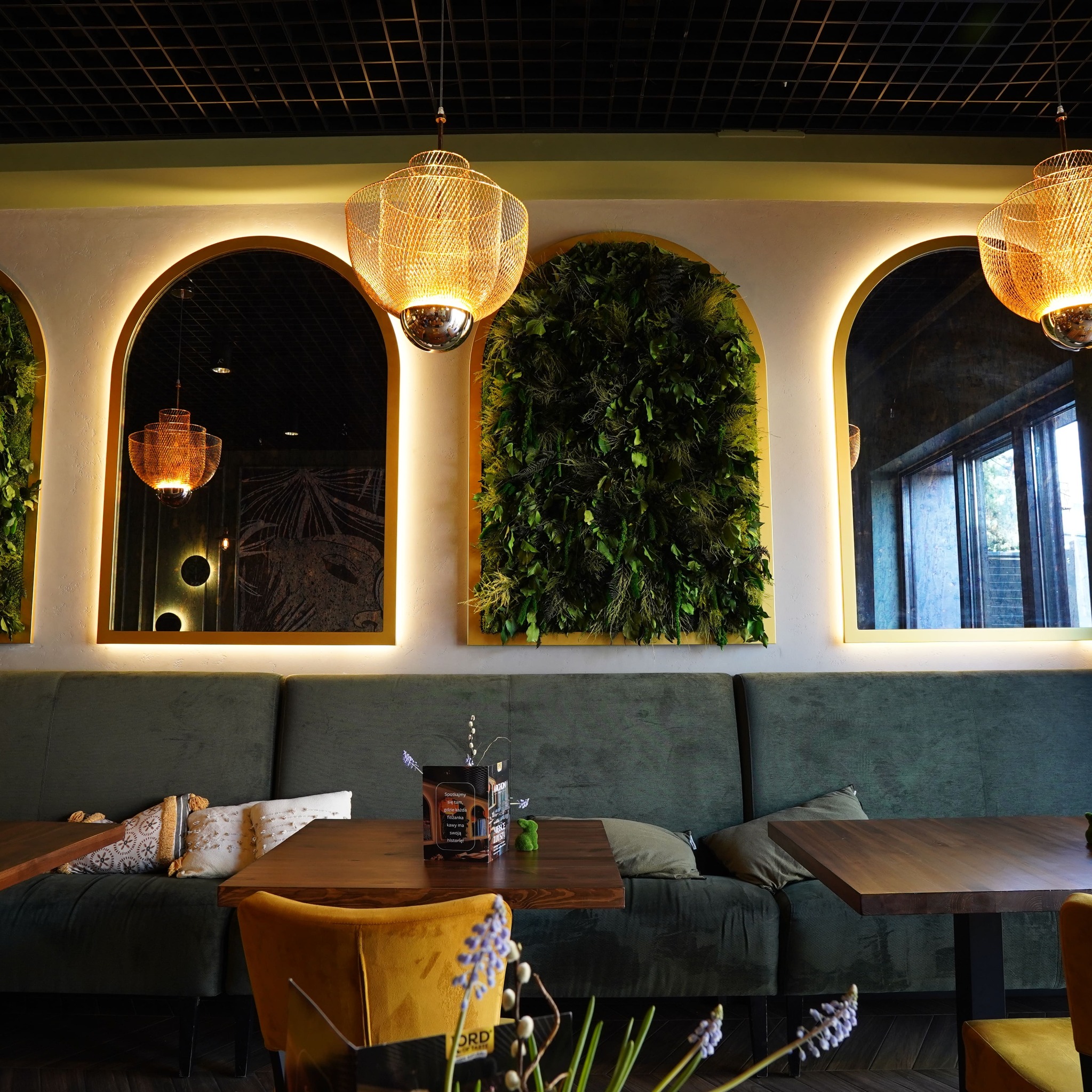 Stylowe wnętrze restauracji z zieloną ścianą, lustrami w złotych ramach i dekoracyjnymi lampami. Wygodne sofy i drewniane stoły tworzą przytulną atmosferę.