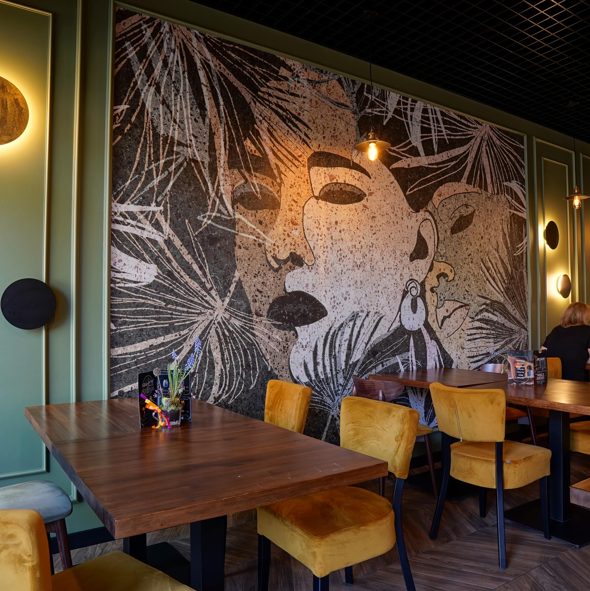 Mozaika z motywem twarzy i roślin na ścianie w lokalu. Oświetlenie punktowe, drewniane stoły i żółte krzesła tworzą przytulną atmosferę.