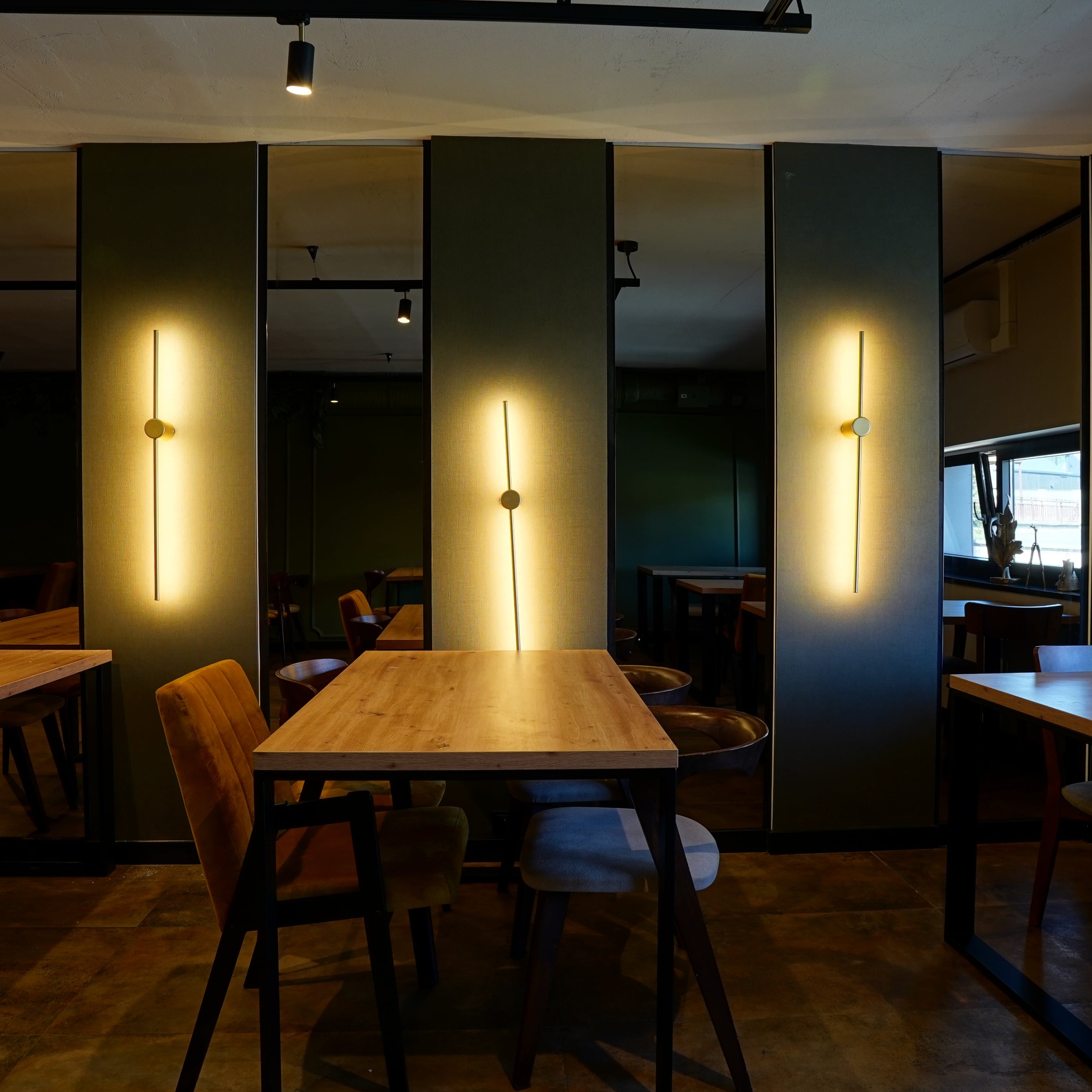 Nowoczesne oświetlenie ścienne w lokalu usługowym. Pionowe lampy z okrągłym akcentem na tle paneli i luster. Widoczne stoły i krzesła w tle.