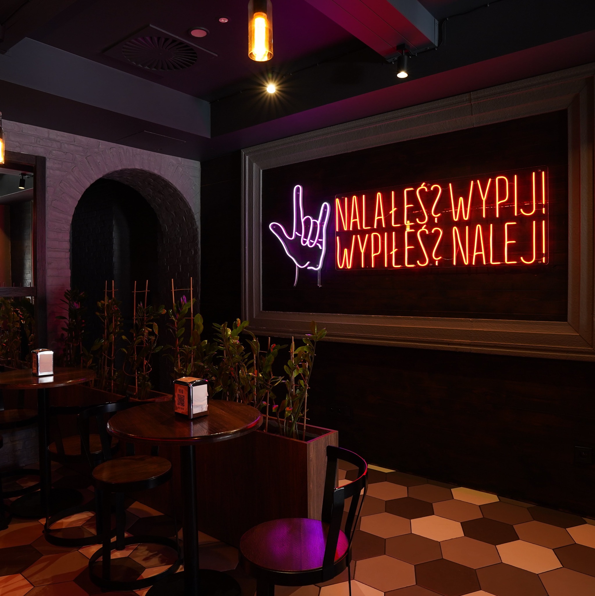Nowoczesne wnętrze baru z neonowym napisem 'Nalałeś? Wypij! Wypiłeś? Nalej!', okrągłymi stolikami i krzesłami. Ciemna kolorystyka, heksagonalna podłoga.