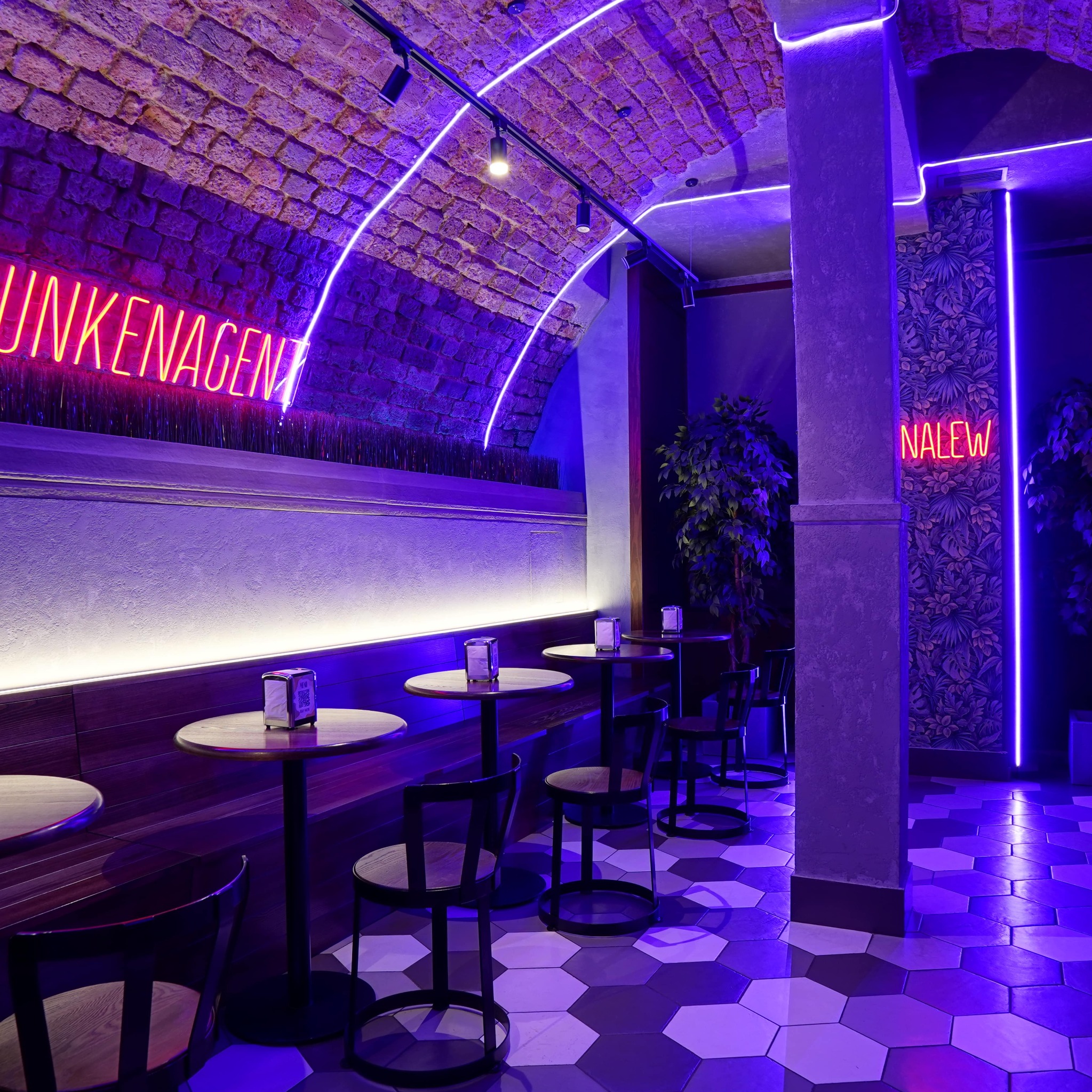 Nowoczesne wnętrze lokalu z ceglanym sklepieniem, neonami i geometryczną podłogą w odcieniach fioletu. Widoczne stoliki i krzesła.