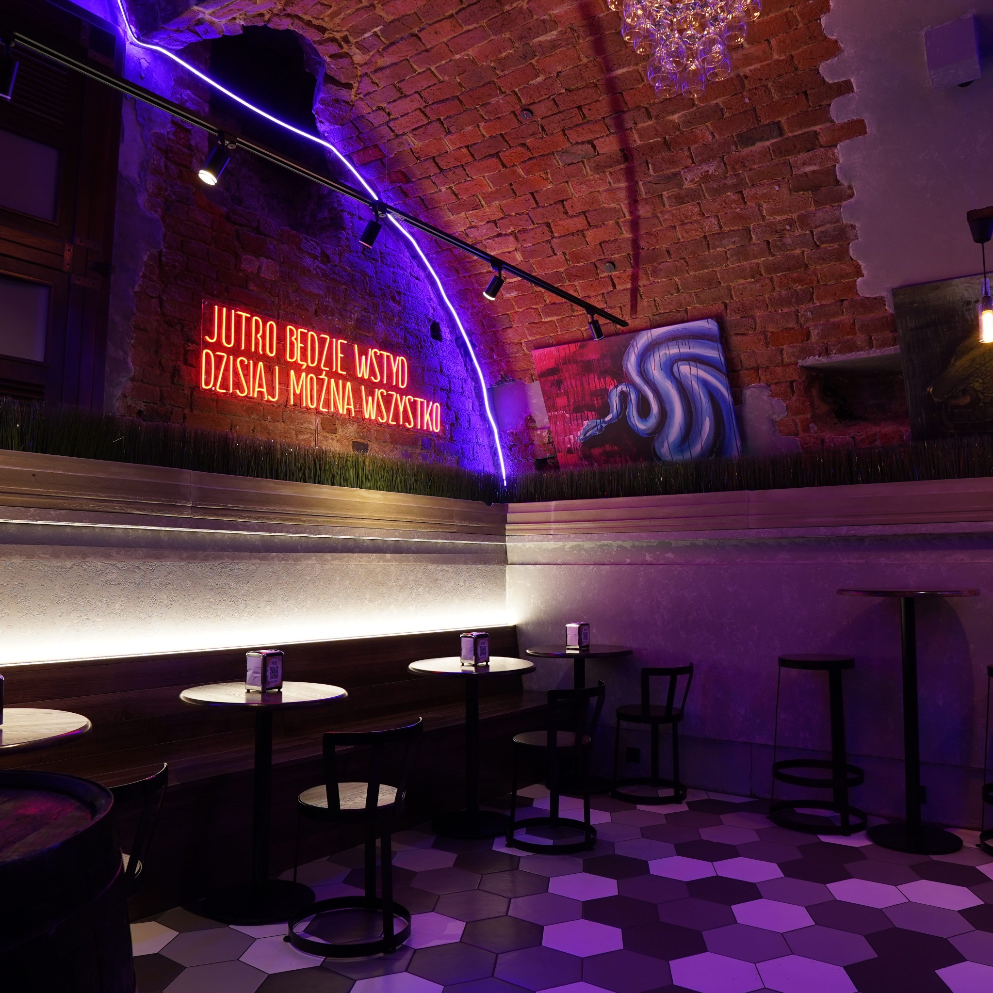 Klimatyczne wnętrze lokalu z ceglanym sklepieniem, neonem i geometryczną posadzką. Oświetlenie punktowe i dekoracyjne. Widoczne stoliki i krzesła barowe.