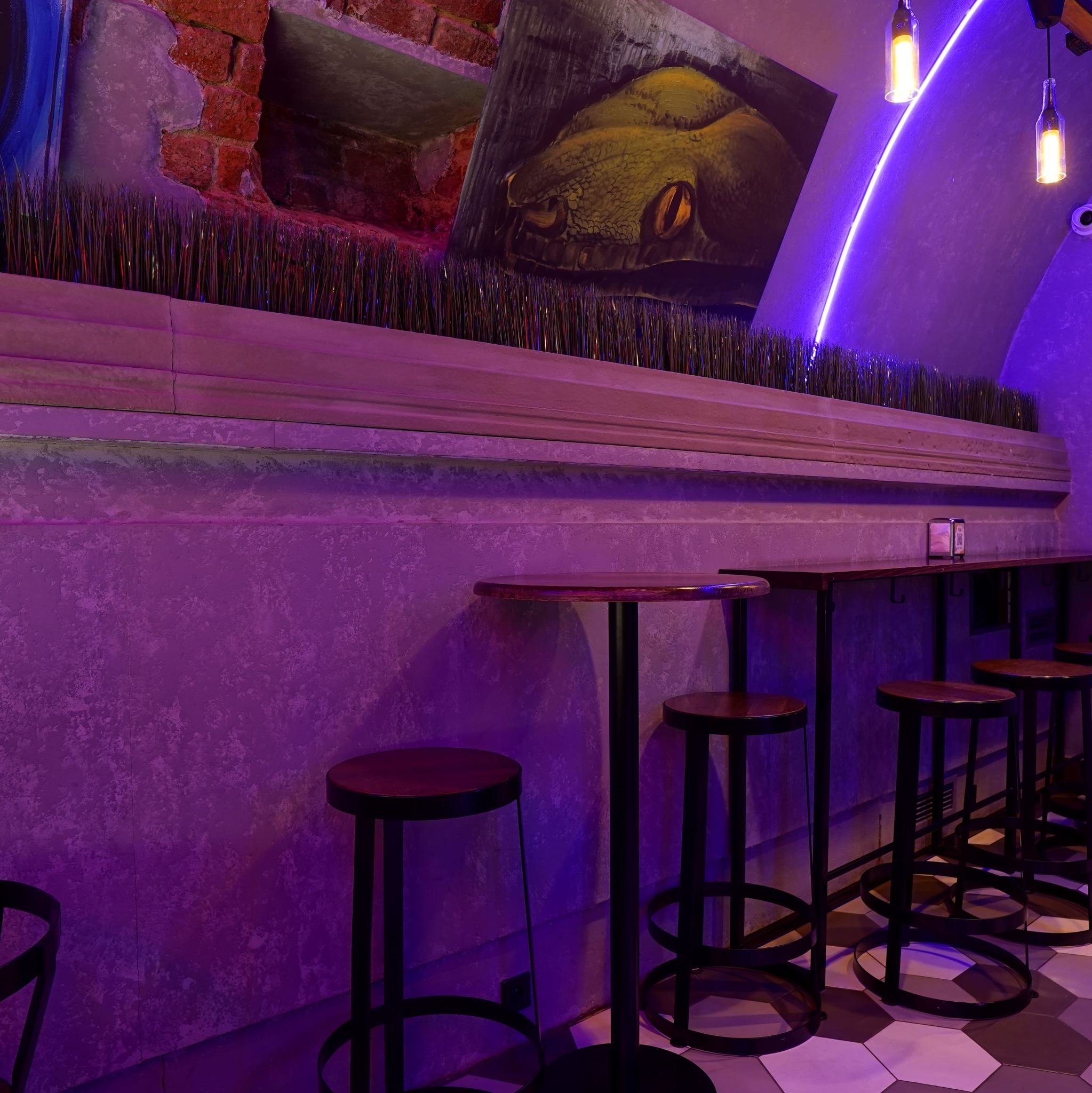 Klimatyczne wnętrze baru z wysokimi stołkami, dekoracyjną ścianą z motywem węża i neonowym oświetleniem w odcieniach fioletu. Nietypowy wystrój.