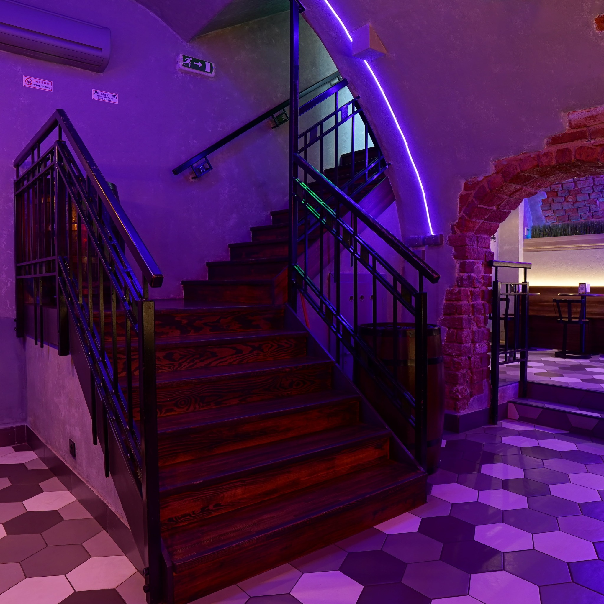 Klatka schodowa z drewnianymi schodami i metalową balustradą, oświetlona fioletowym neonem. Widoczny fragment ceglanego łuku i wzorzysta podłoga.
