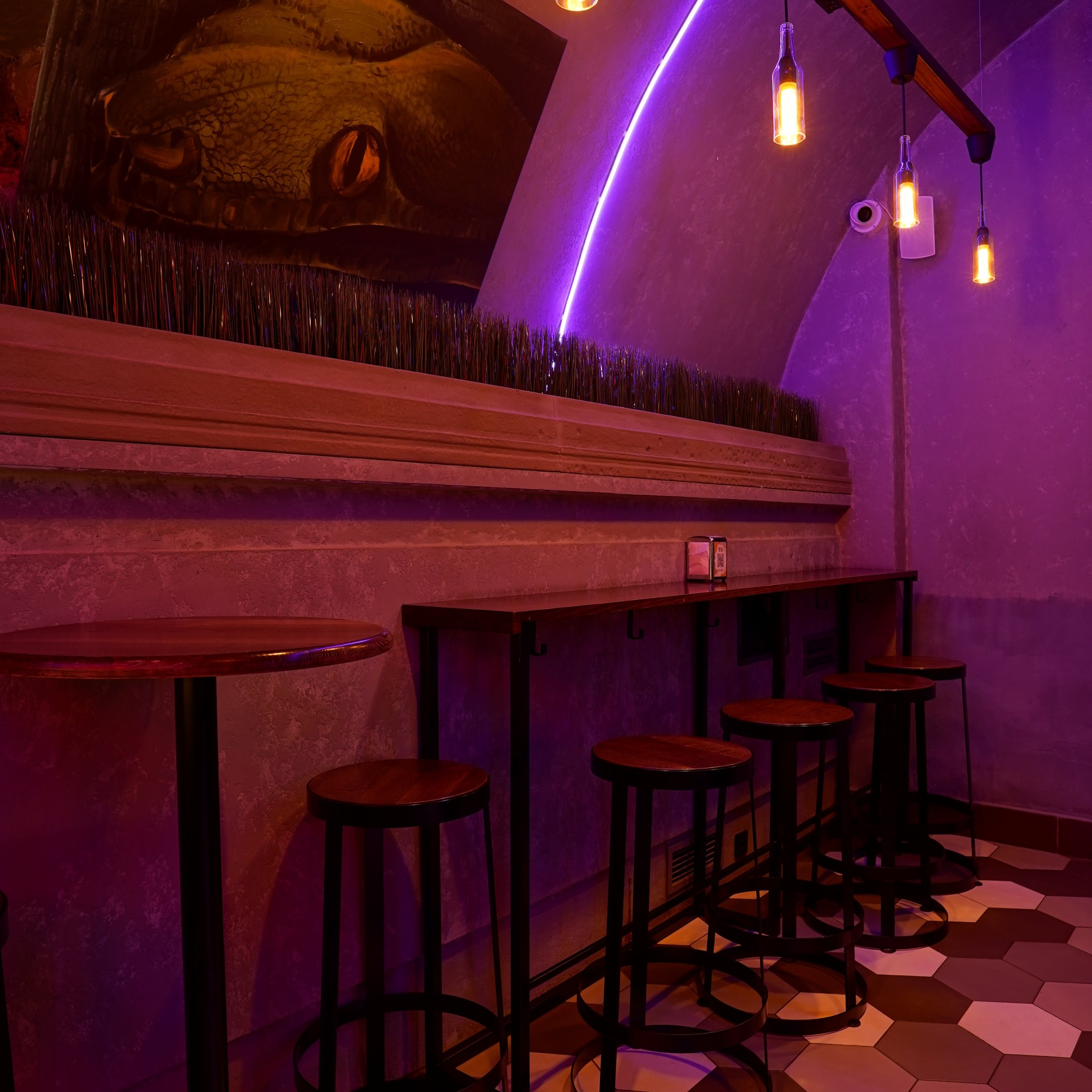 Stylowe wnętrze baru z neonowym oświetleniem i muralem węża. Wysokie stołki barowe przy blacie, heksagonalna podłoga. Unikalny design z akcentem na detale.