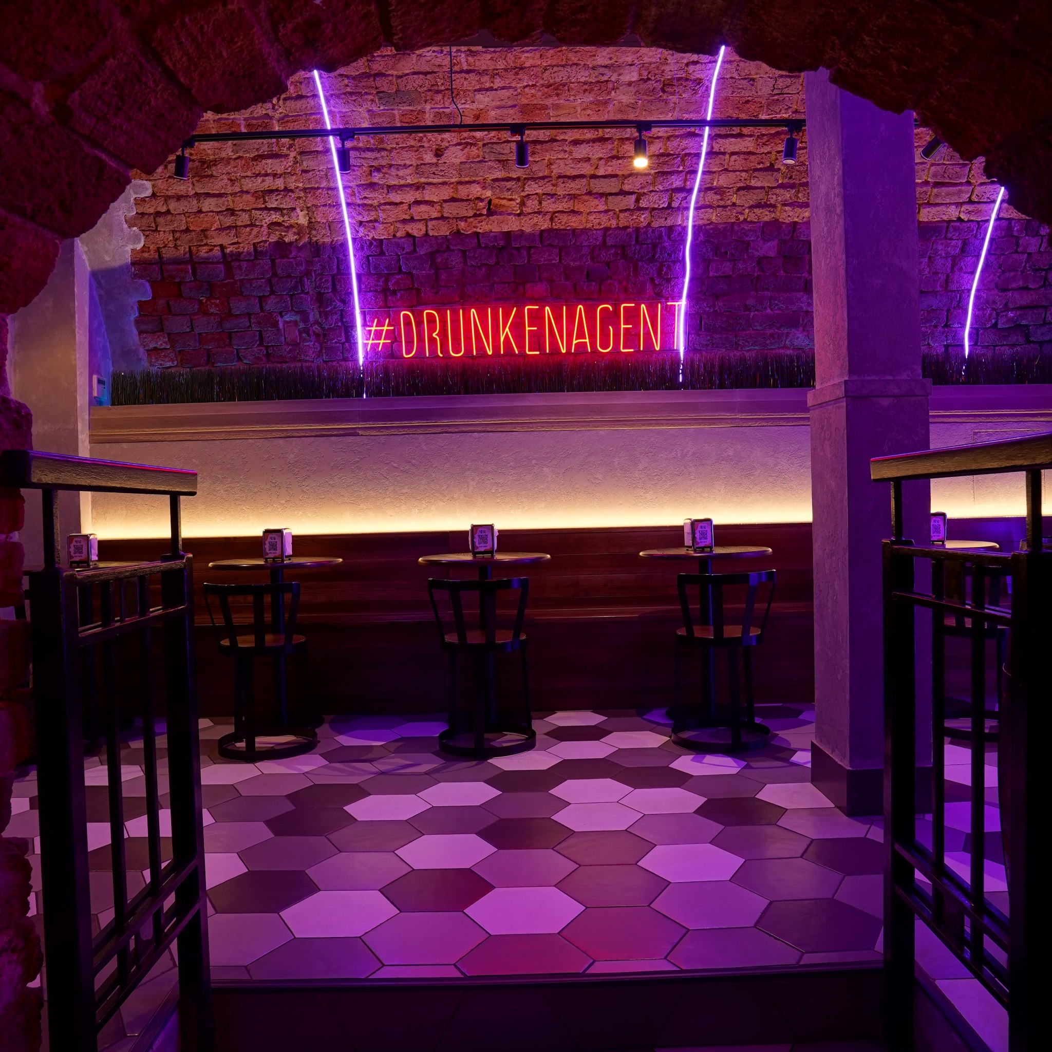Klimatyczne wnętrze baru z ceglaną ścianą, neonem '#DRUNKENAGENT' i geometryczną posadzką w odcieniach fioletu. Widoczne stoliki i krzesła, oświetlenie punktowe.
