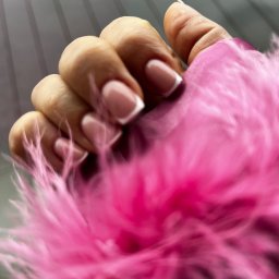 Mariana - Dłoń z perfekcyjnym manicure francuskim na tle różowego boa z piór. Delikatne, jasne paznokcie z białymi końcówkami. Estetyczny i zadbany wygląd.
