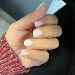 Mariana - Dłoń z białym manicure francuskim na krótkich, kwadratowych paznokciach, widoczna na tle czarnego materiału i fragmentu swetra w odcieniach czerwieni i beżu.