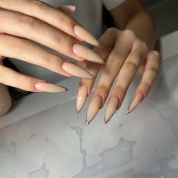 Mariana - Dłonie z beżowymi, błyszczącymi paznokciami w kształcie sztyletu, na jasnym tle. Widoczne detale manicure, precyzyjne wykończenie.