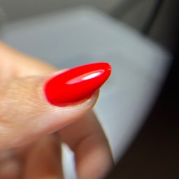 Mariana - Błyszczący, czerwony paznokieć w zbliżeniu, z delikatnym połyskiem i owalnym kształtem, na tle rozmytego, jasnego tła. Widoczna struktura skóry palca.