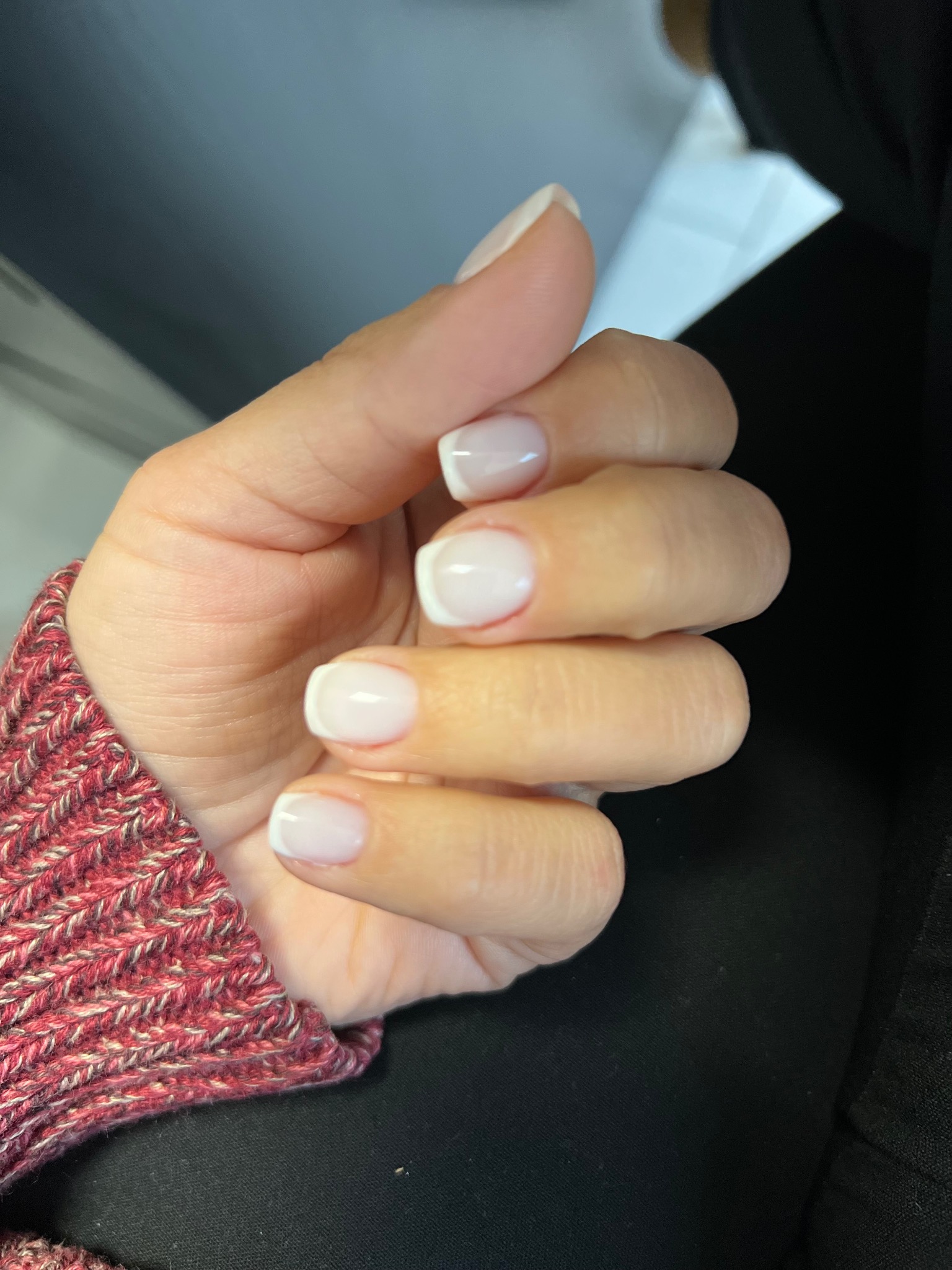 Dłoń z białym manicure francuskim na krótkich, kwadratowych paznokciach, widoczna na tle czarnego materiału i fragmentu swetra w odcieniach czerwieni i beżu.