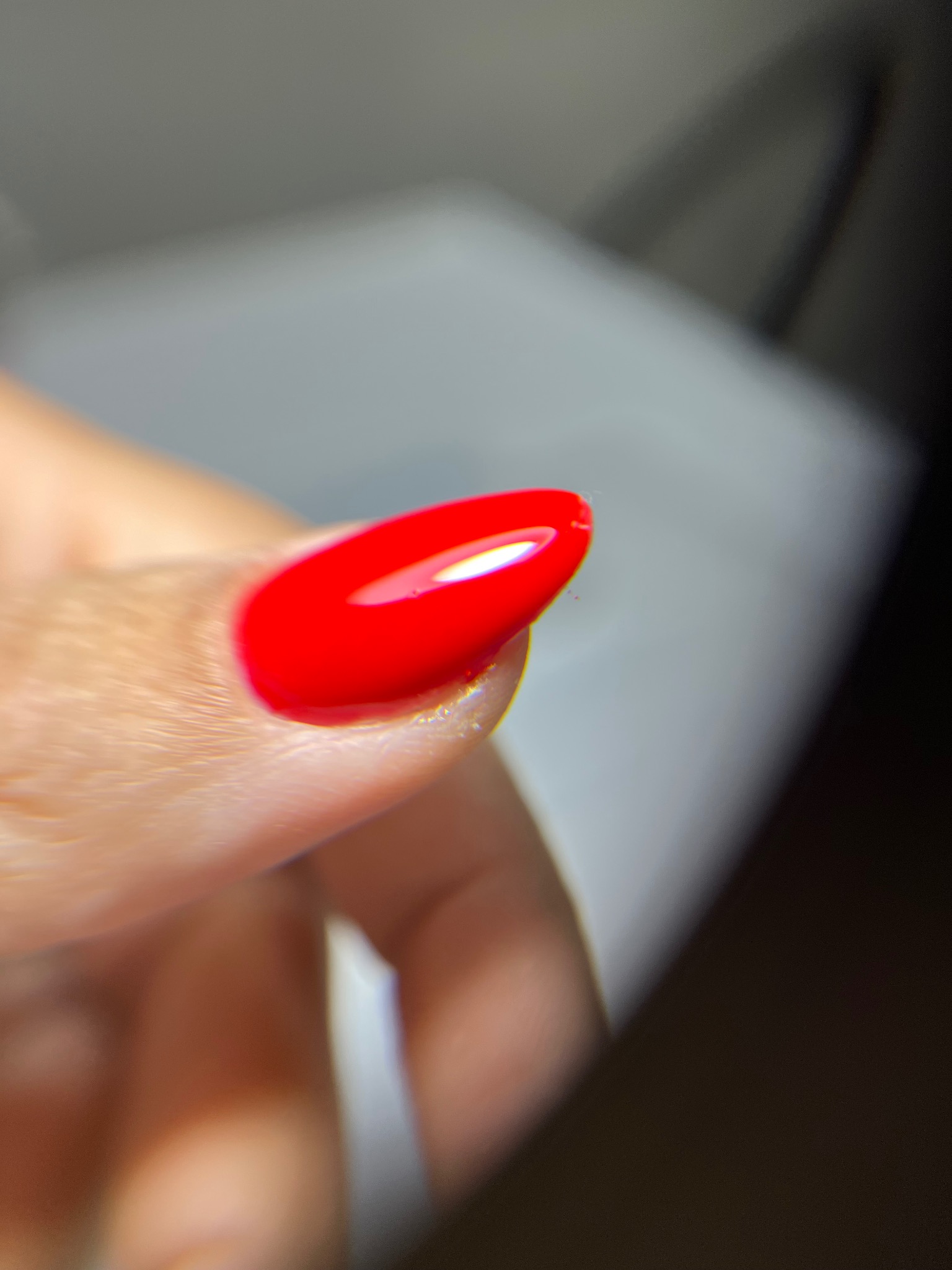 Błyszczący, czerwony paznokieć w zbliżeniu, z delikatnym połyskiem i owalnym kształtem, na tle rozmytego, jasnego tła. Widoczna struktura skóry palca.