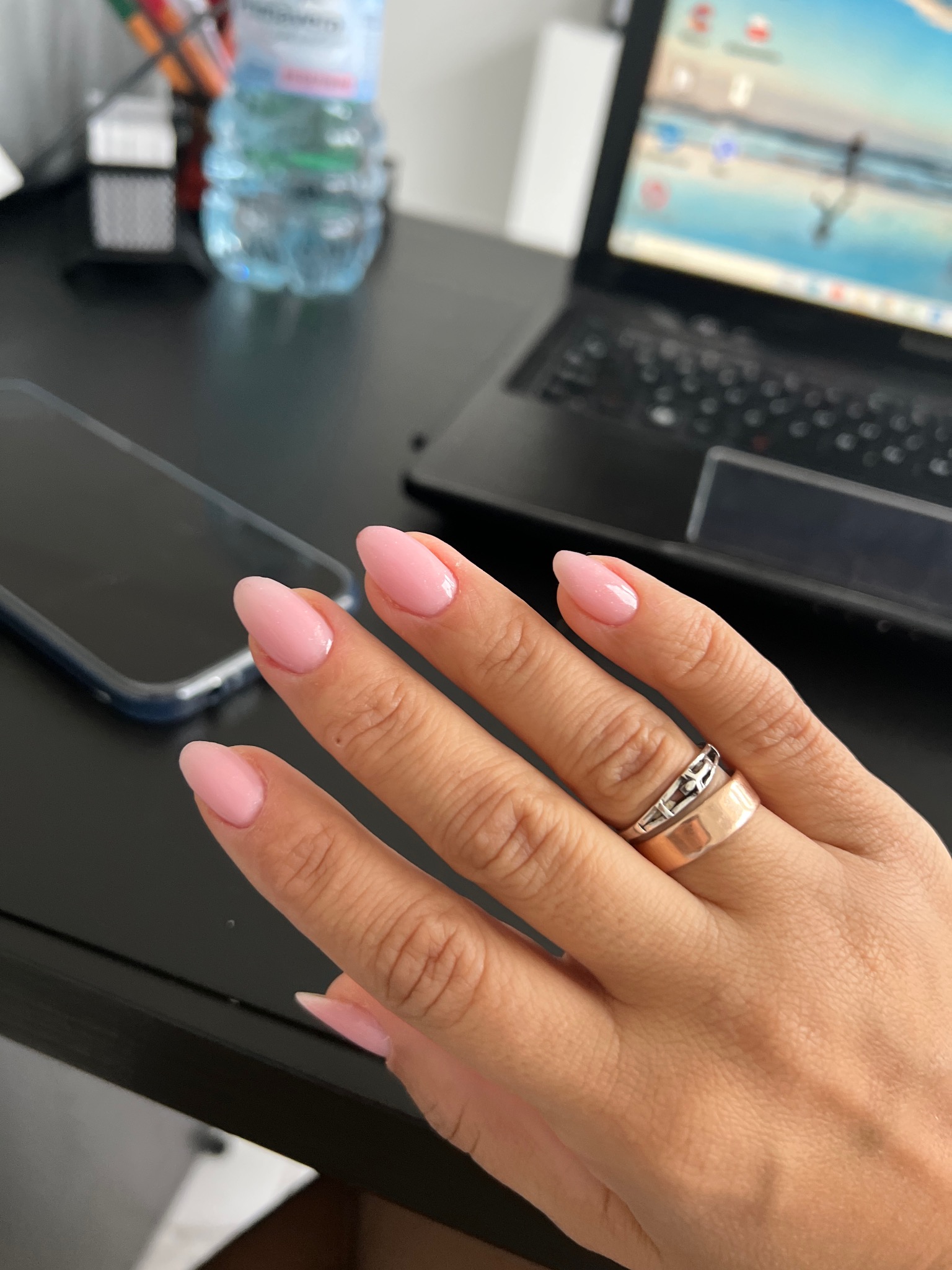 Ręka z delikatnym, różowym manicure hybrydowym na tle biurka z laptopem i telefonem. Pierścionek na palcu serdecznym. Estetyczne, zadbane paznokcie.
