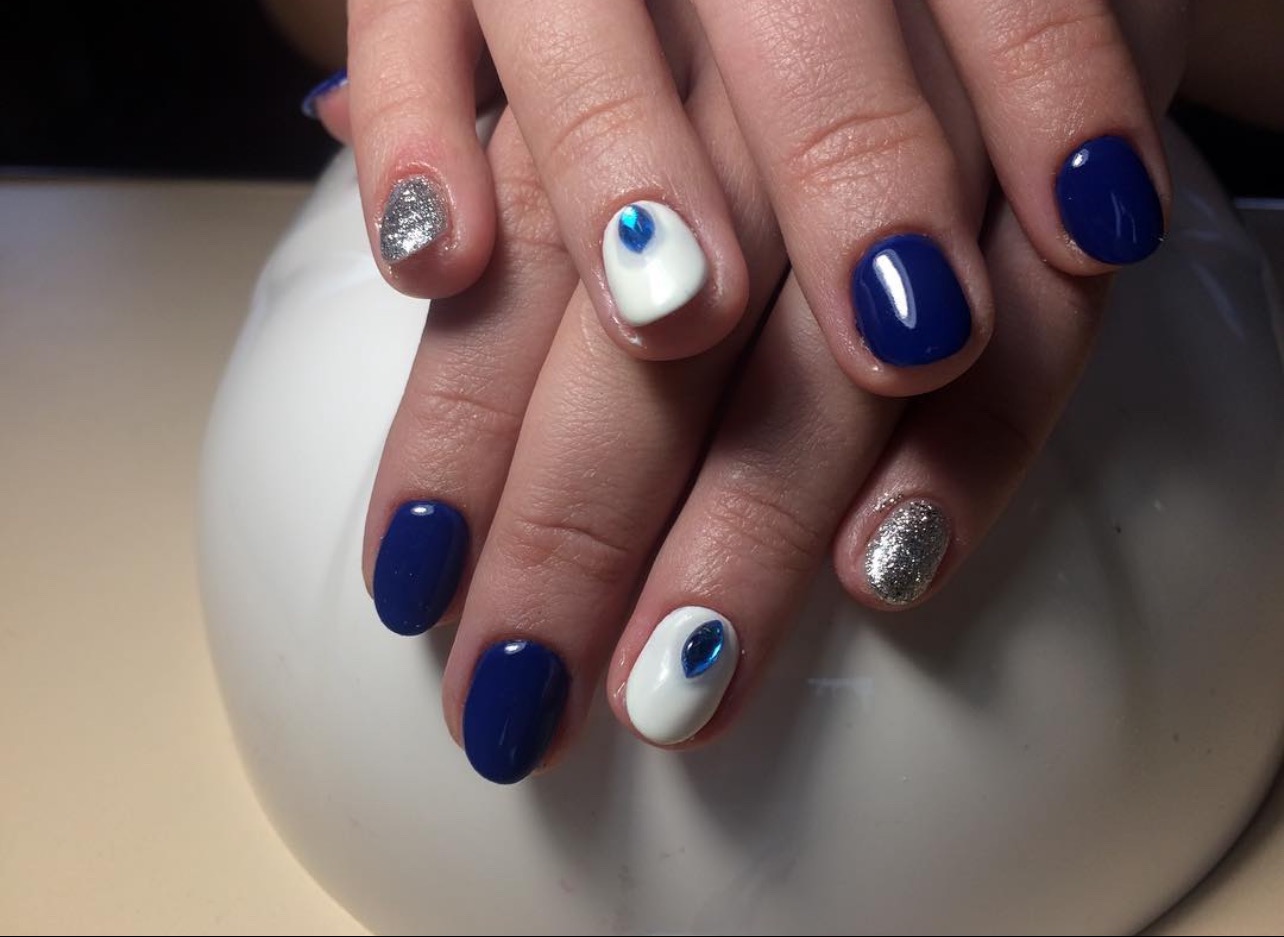 Dłonie z manicure hybrydowym: granatowe, białe z niebieskim oczkiem i srebrne brokatowe paznokcie. Elegancki i nowoczesny design.