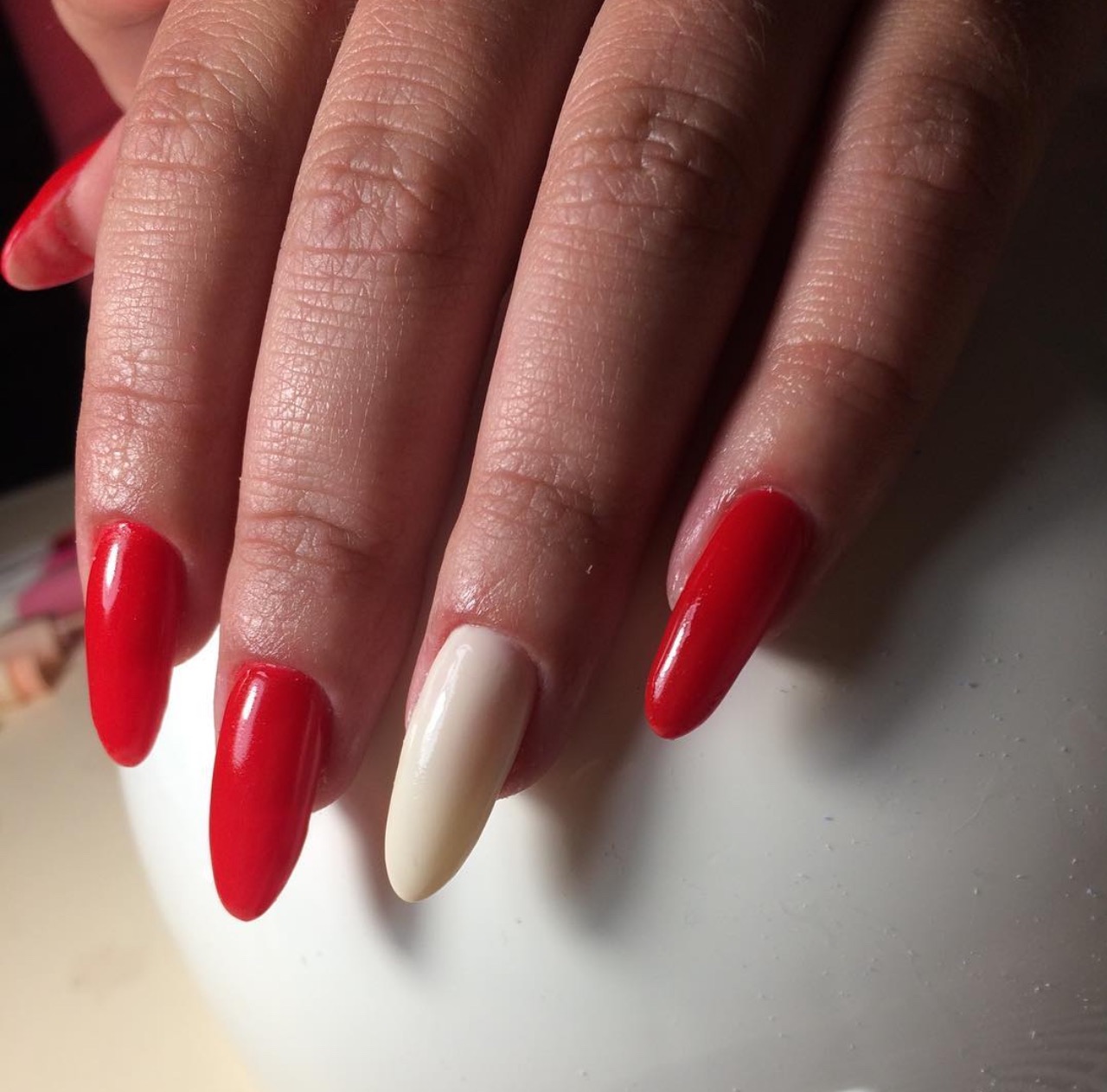 Dłoń z pomalowanymi paznokciami: trzy czerwone, jeden kremowy. Manicure hybrydowy z błyszczącym wykończeniem, widoczne detale skóry dłoni.